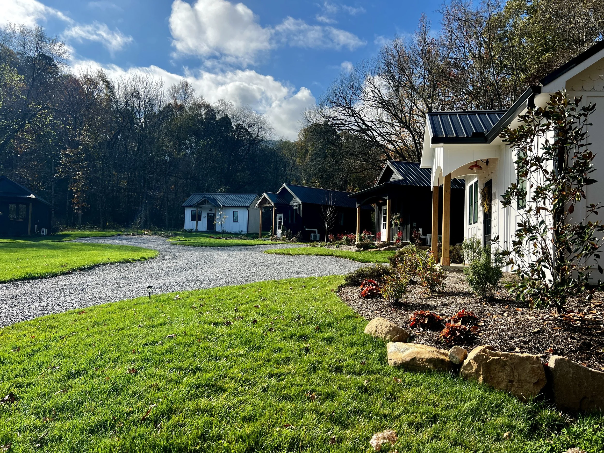 Small cottages along a gravel path with green lawns and trees in the background under a blue sky with clouds.