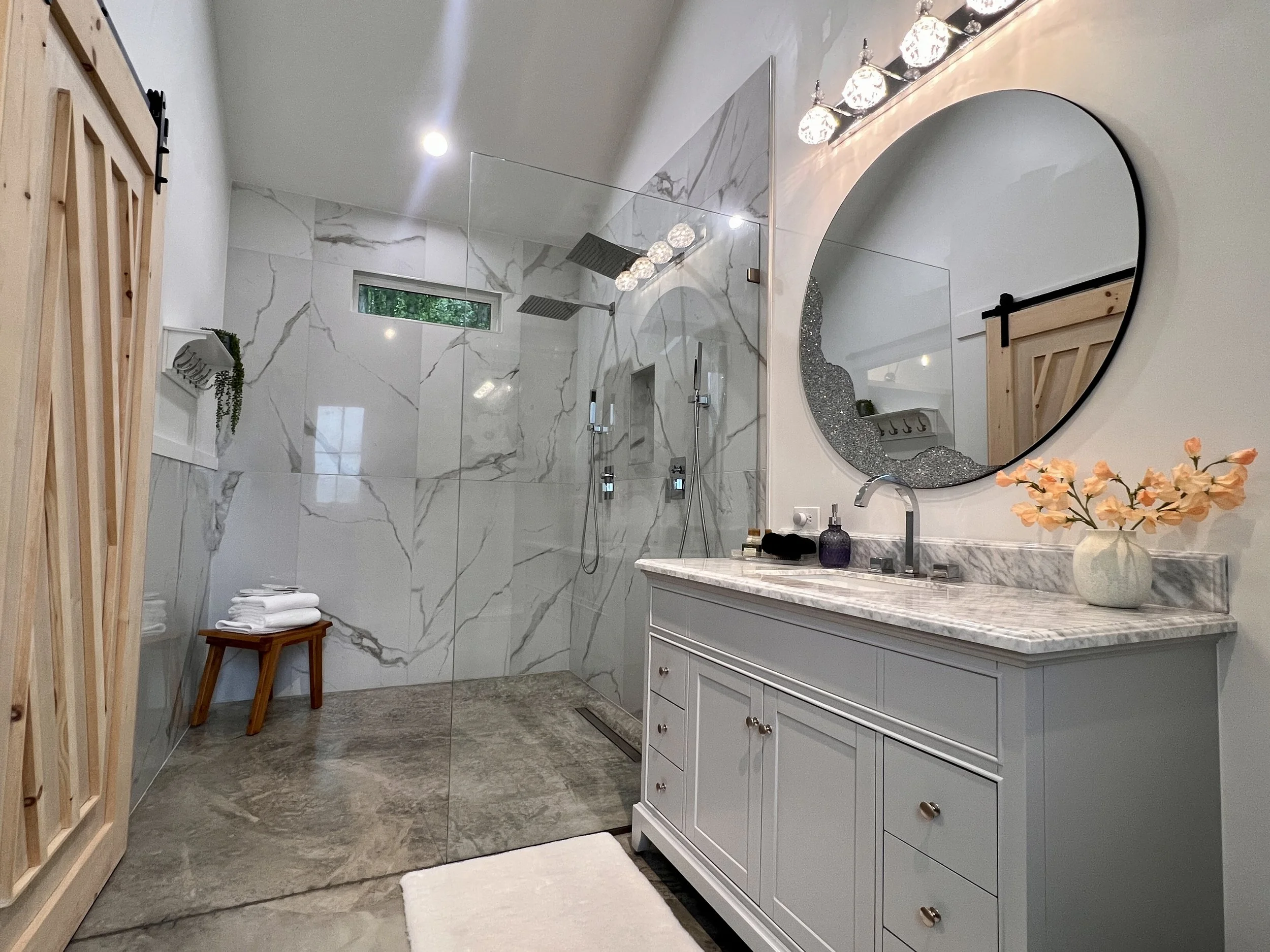 Modern bathroom with glass shower, round mirror, marble countertop, light fixture, wooden barn door, and decorative plants.