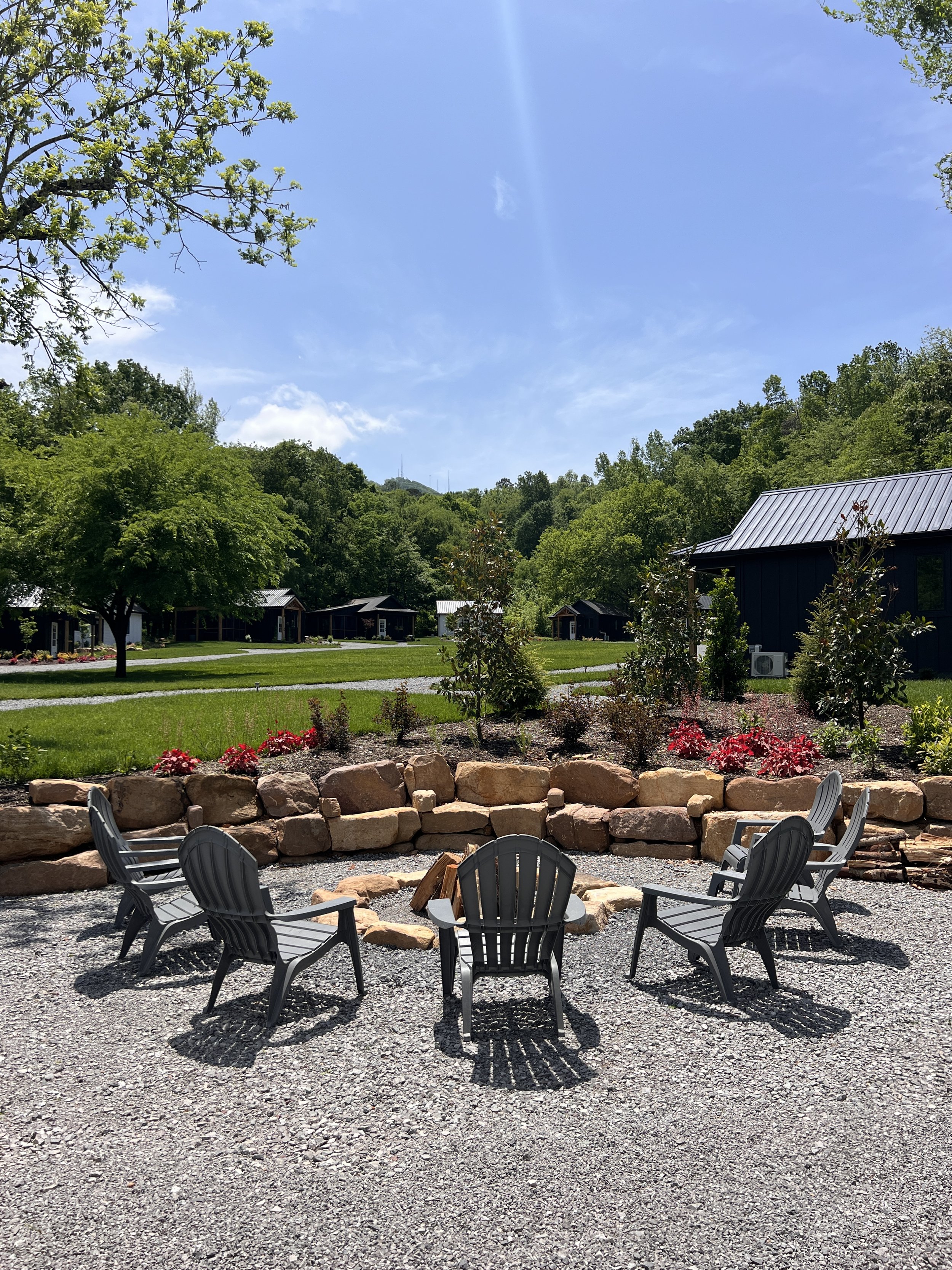 Outdoor seating area with Adirondack chairs around a fire pit, surrounded by trees and a stone wall on a sunny day.