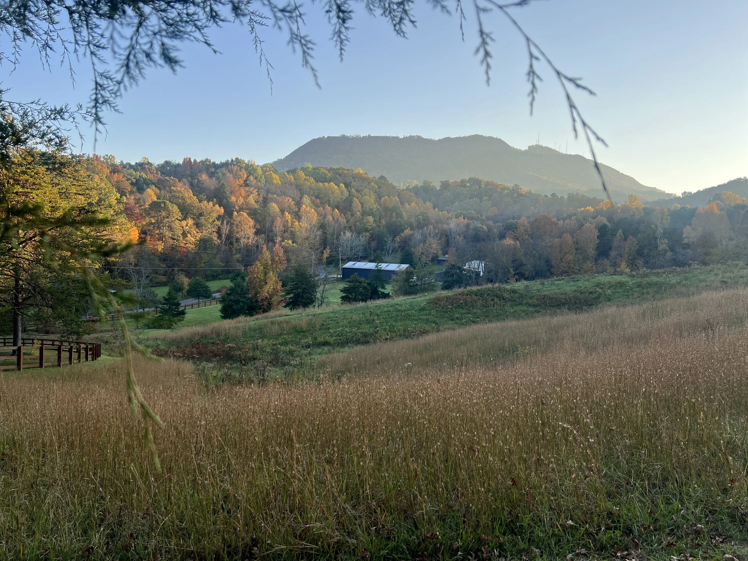 Countryside landscape with rolling hills, grassy field, and autumn trees, with a house and barn in the distance against a backdrop of mountains under a clear sky.