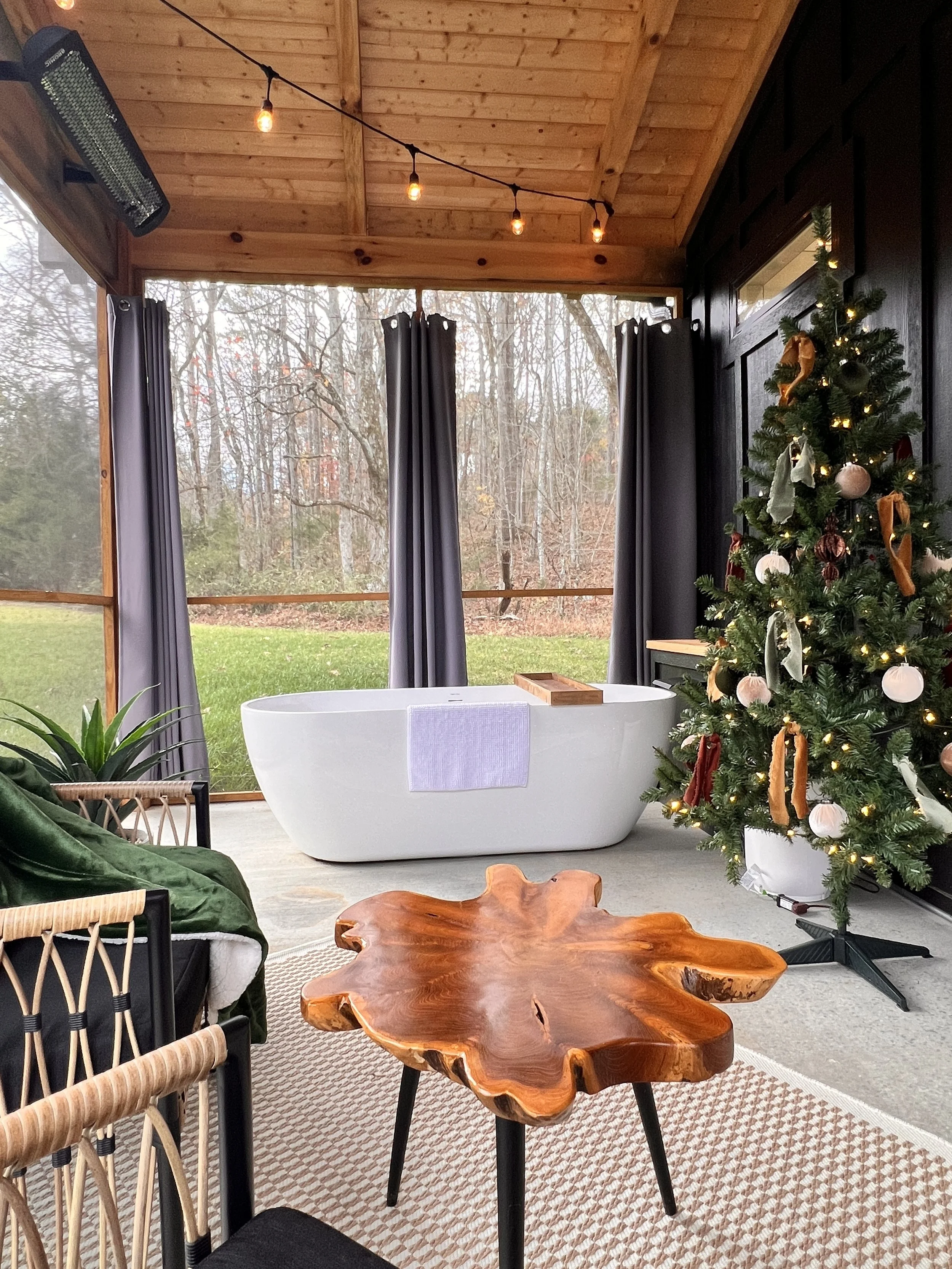 Outdoor setting with a freestanding bathtub, a Christmas tree with ornaments, a wooden coffee table, wicker chairs, and string lights under a wooden roof.