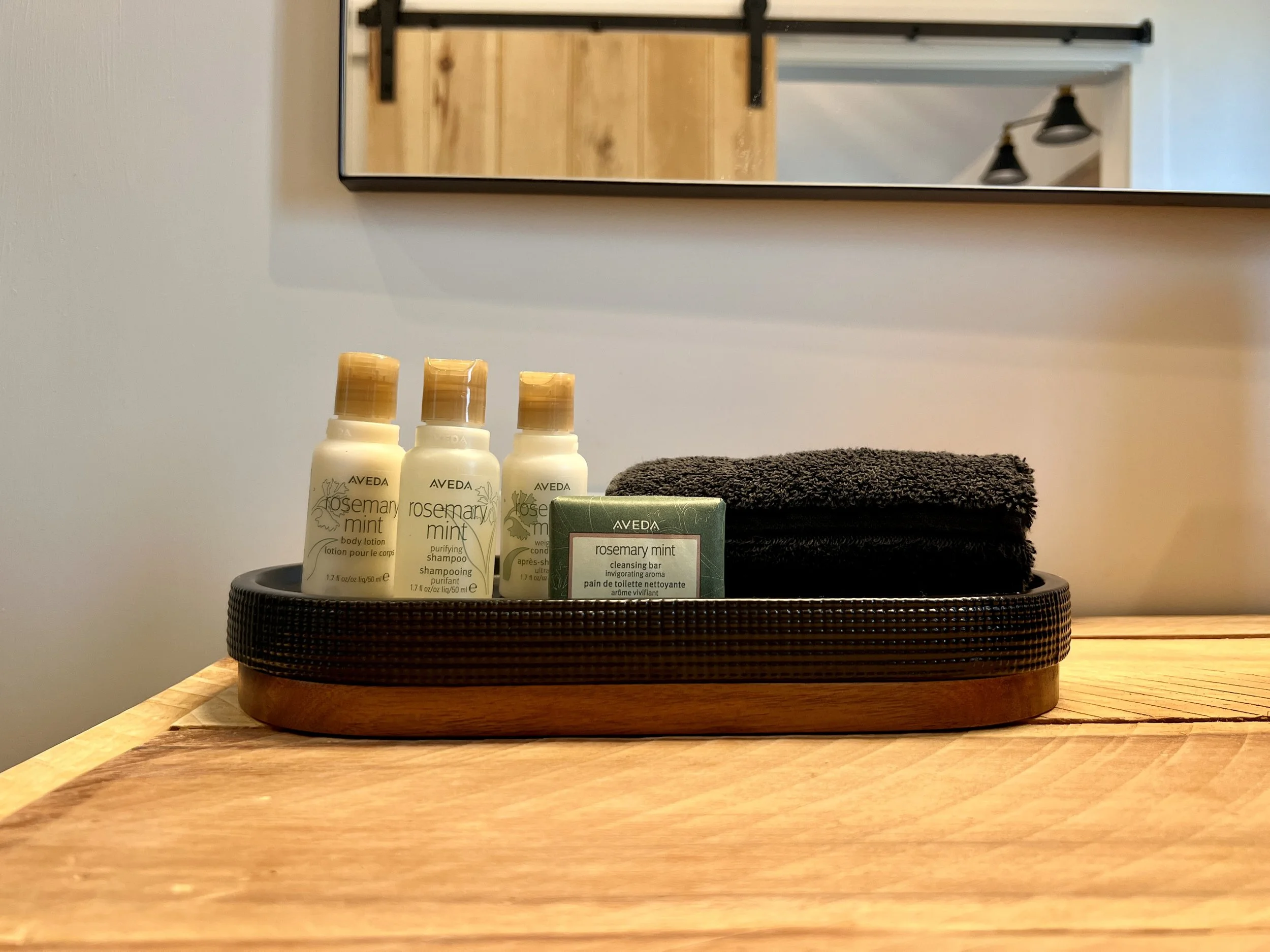 A wooden tray on a wooden surface containing Aveda Rosemary Mint toiletries, including body lotion, shampoo, conditioner, a cleansing bar, and two folded black towels. A mirror and interior lights are visible in the background.