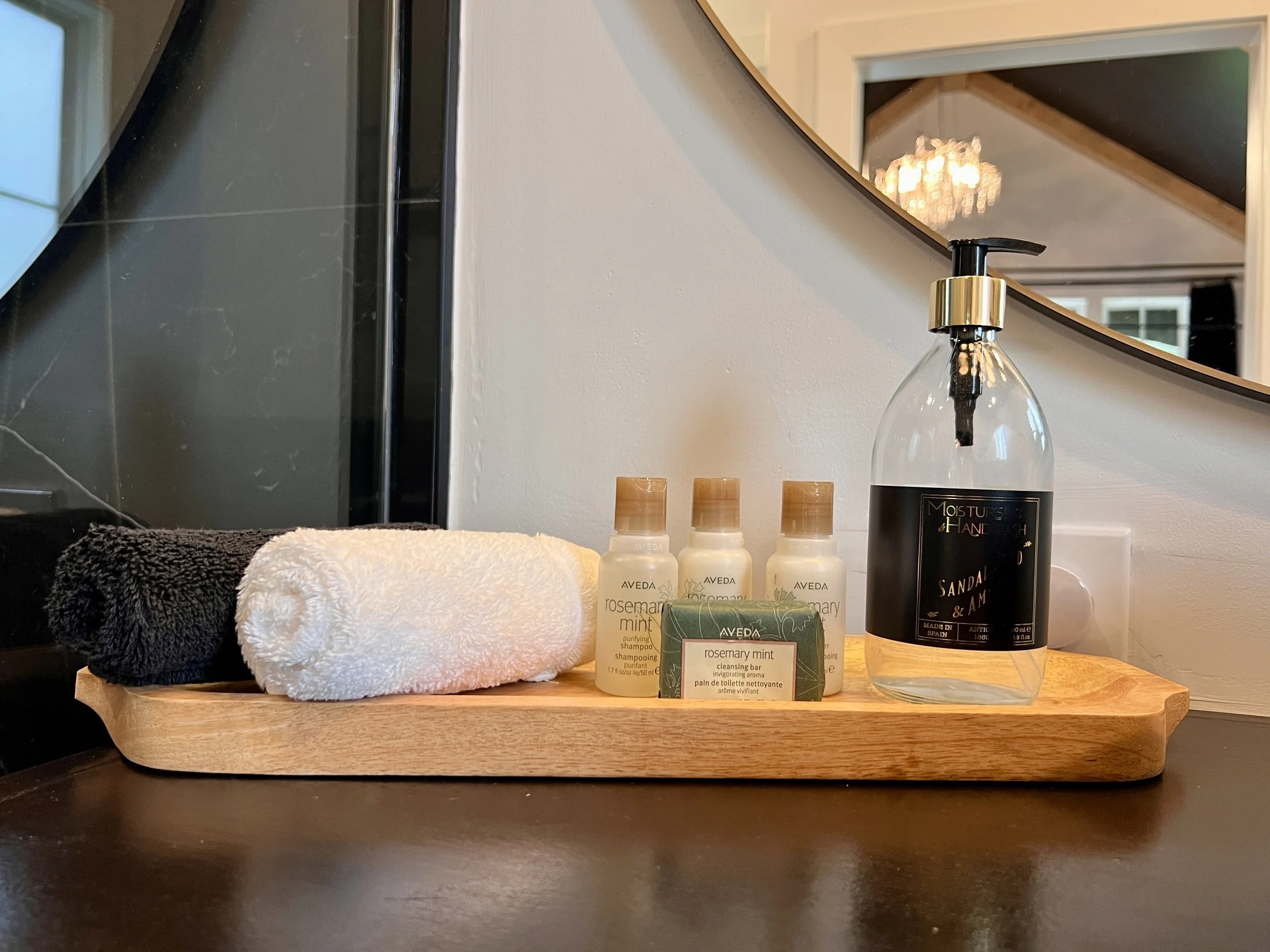 Bathroom amenities on a wooden tray with rolled towels, shampoo, soap, and a hand wash bottle near a mirror.
