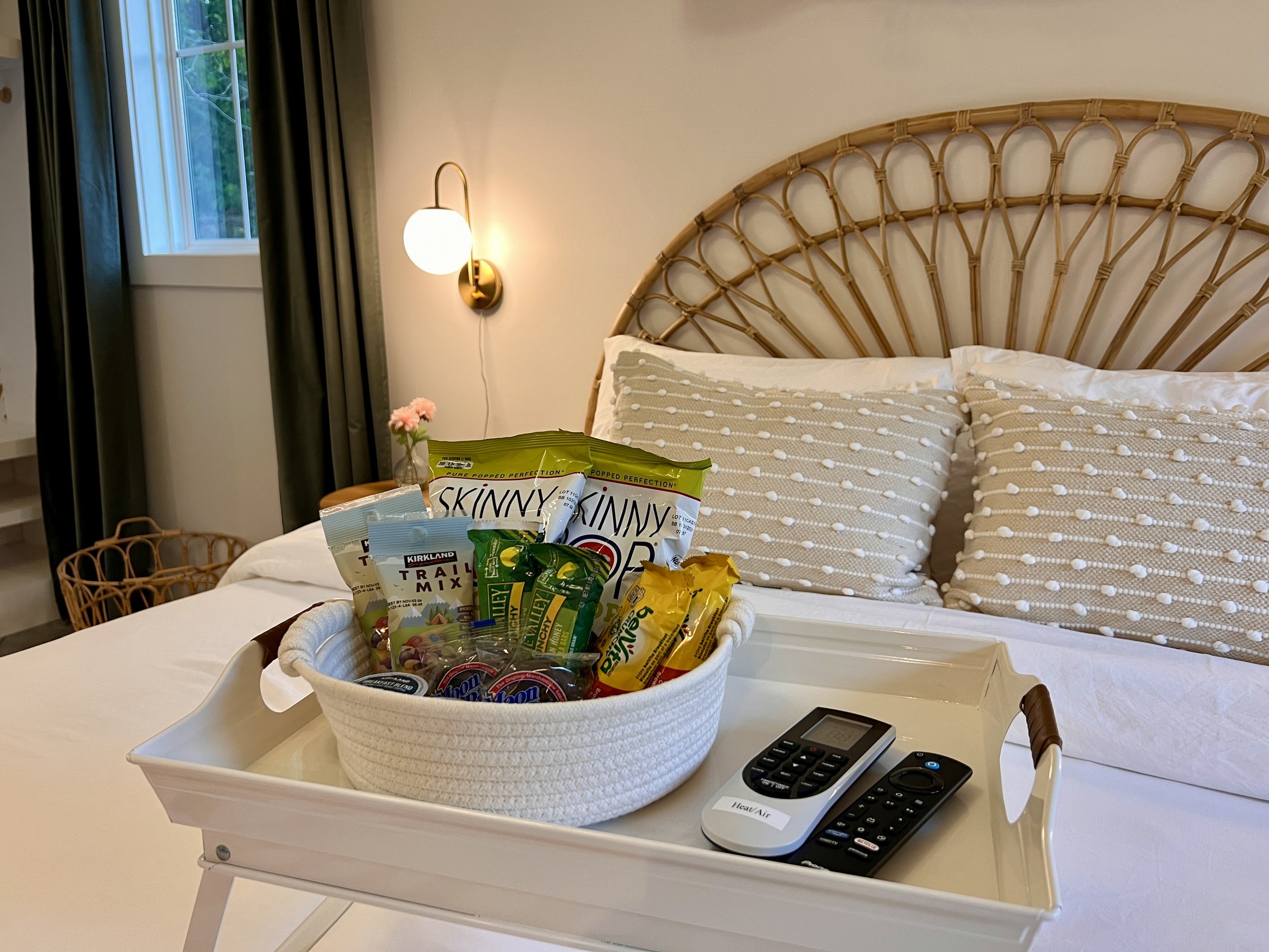 Bedroom with a white bed, wicker headboard, basket of snacks, and two remote controls on a tray. Wall lamp and window in the background.