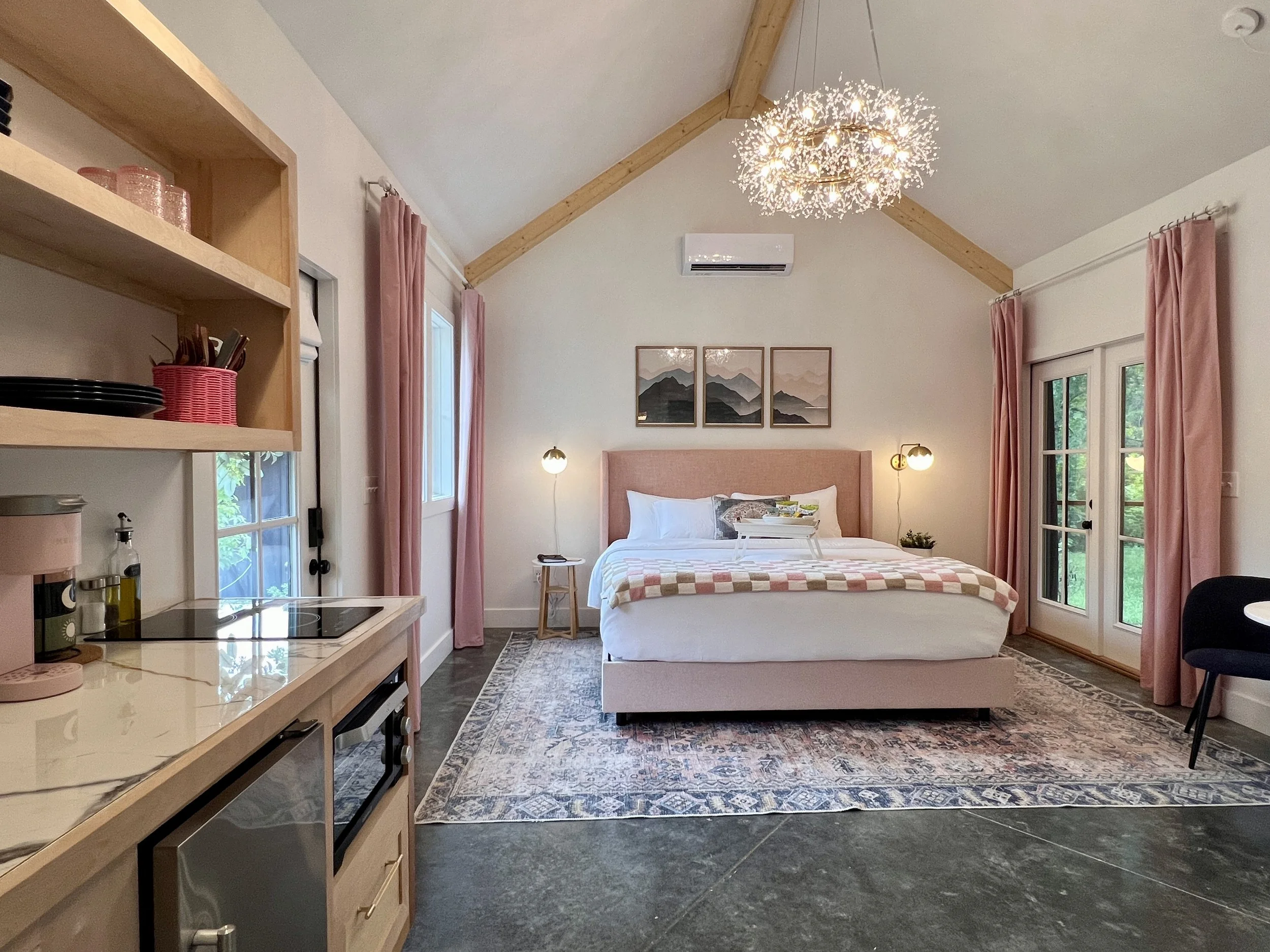 Cozy bedroom with a modern design, featuring a bed with decorative pillows, a geometric chandelier, artwork, and an adjoining kitchen area with a marble countertop.