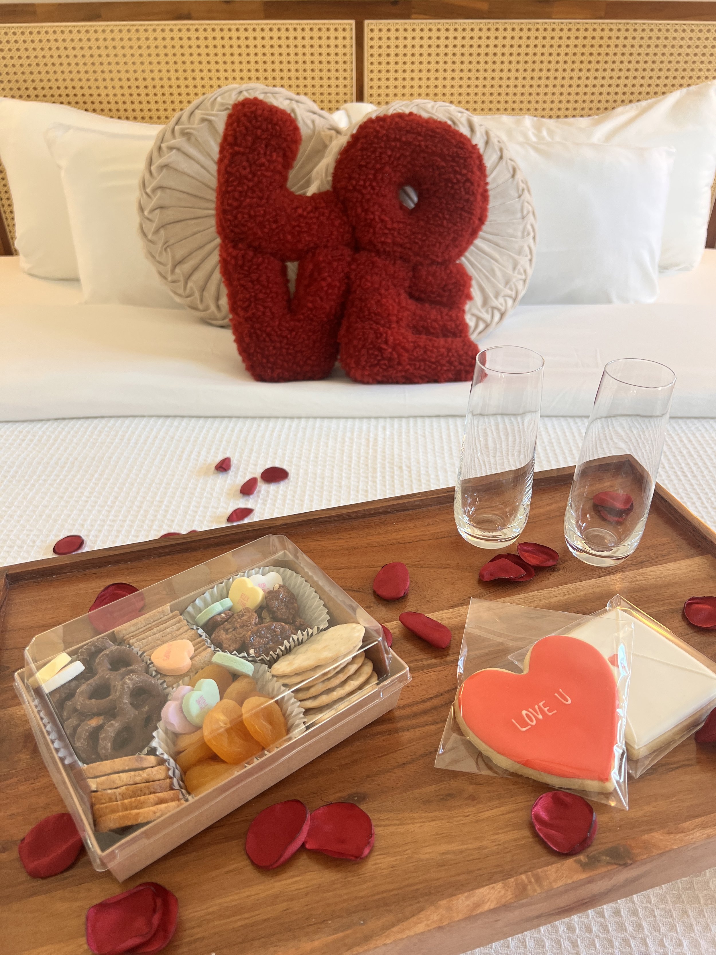 A cozy bedroom setting featuring a wooden tray on a bed, adorned with red rose petals. The tray holds a box of assorted snacks, including pretzels, crackers, and conversation heart candies. Two sealed heart-shaped cookies, one with "LOVE U" written o