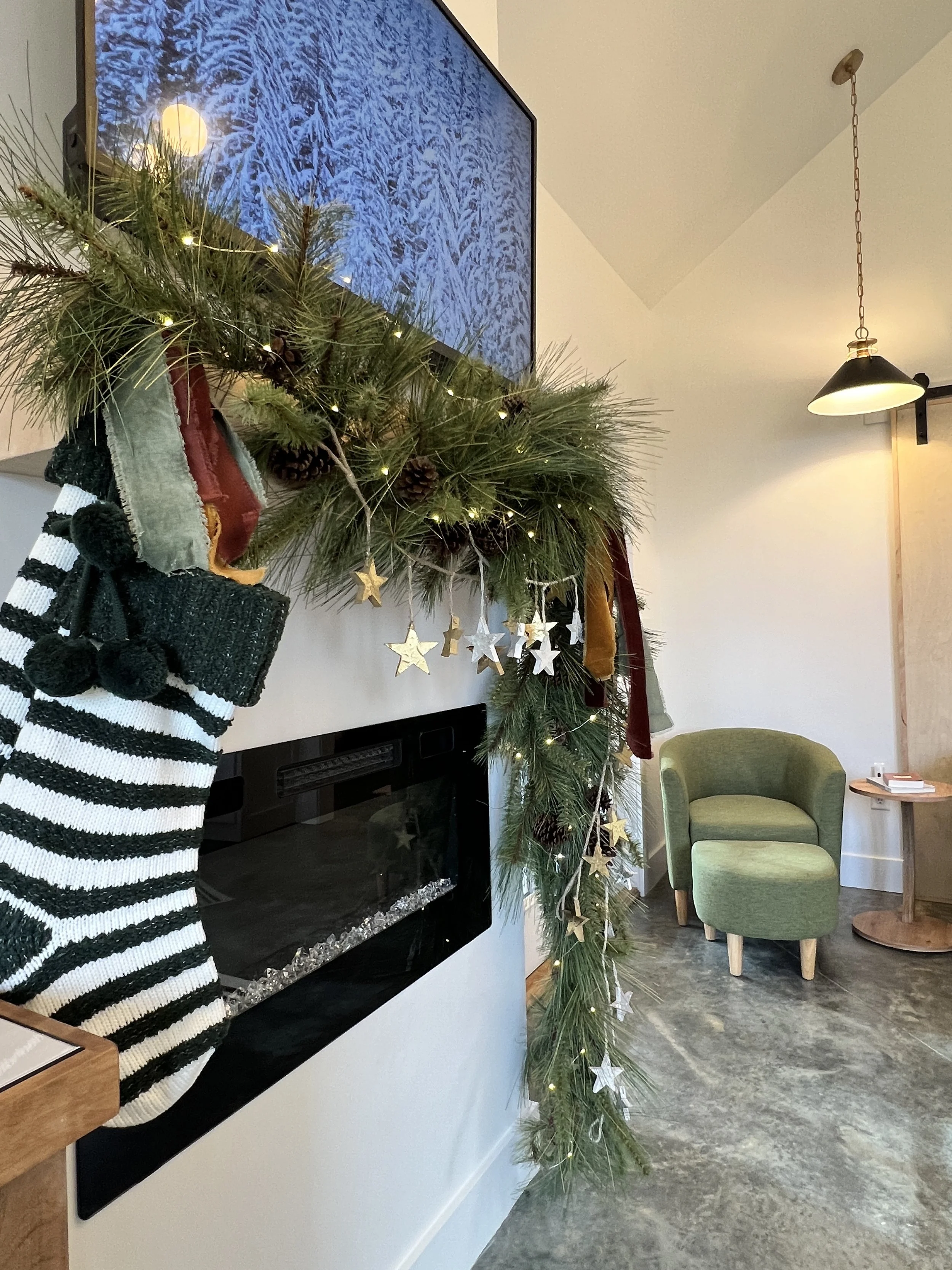 A cozy room with a modern fireplace under a TV screen displaying a snowy forest scene. Greenery garlands with small decorative lights and stars, along with striped Christmas stockings, adorn the fireplace. A green armchair and ottoman sit on a polish