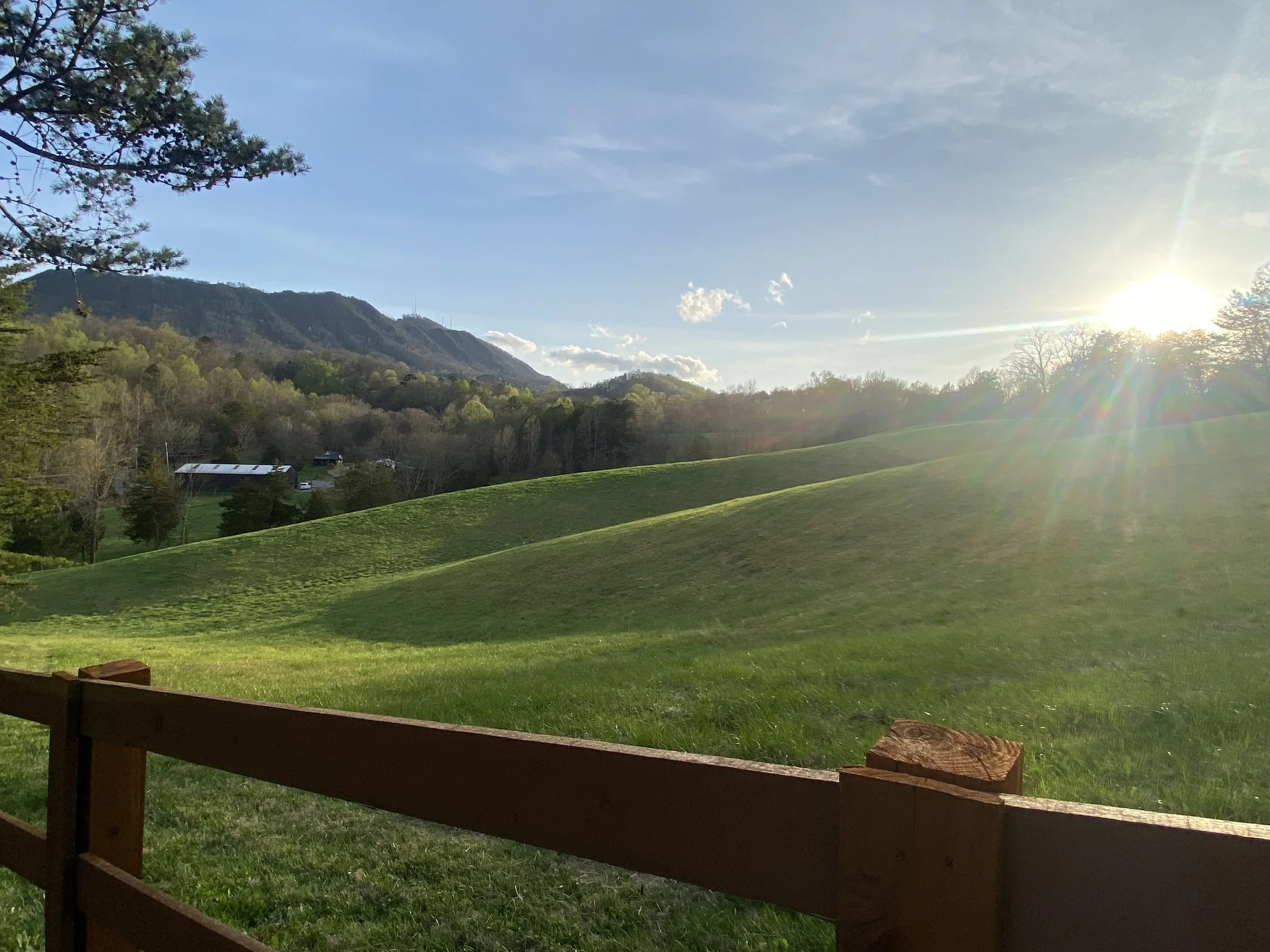 Rolling green hills under a blue sky with the sun shining, a wooden fence in the foreground, trees and mountains in the background.