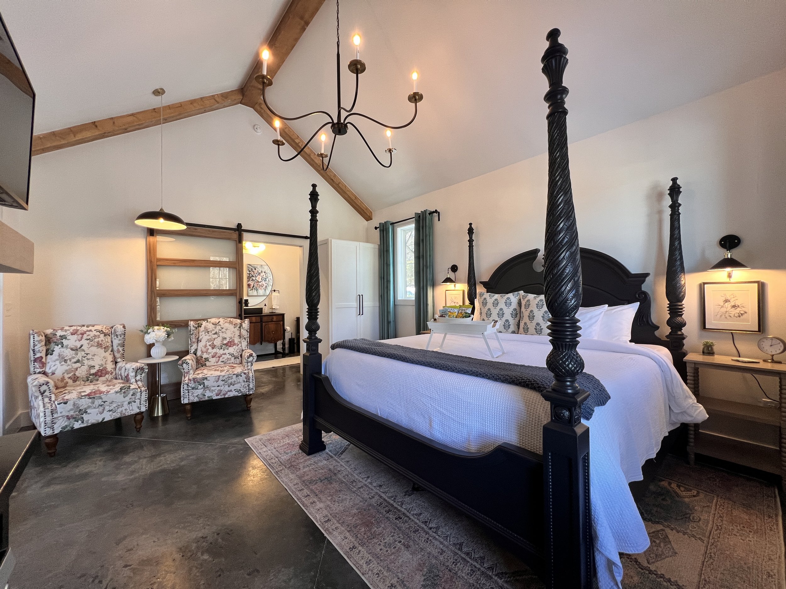 A modern bedroom with a four-poster bed, floral armchairs, and a chandelier. The room features high ceilings with exposed wooden beams, a sliding wooden door, and a mix of contemporary and rustic decor elements. A cozy atmosphere is created by the li