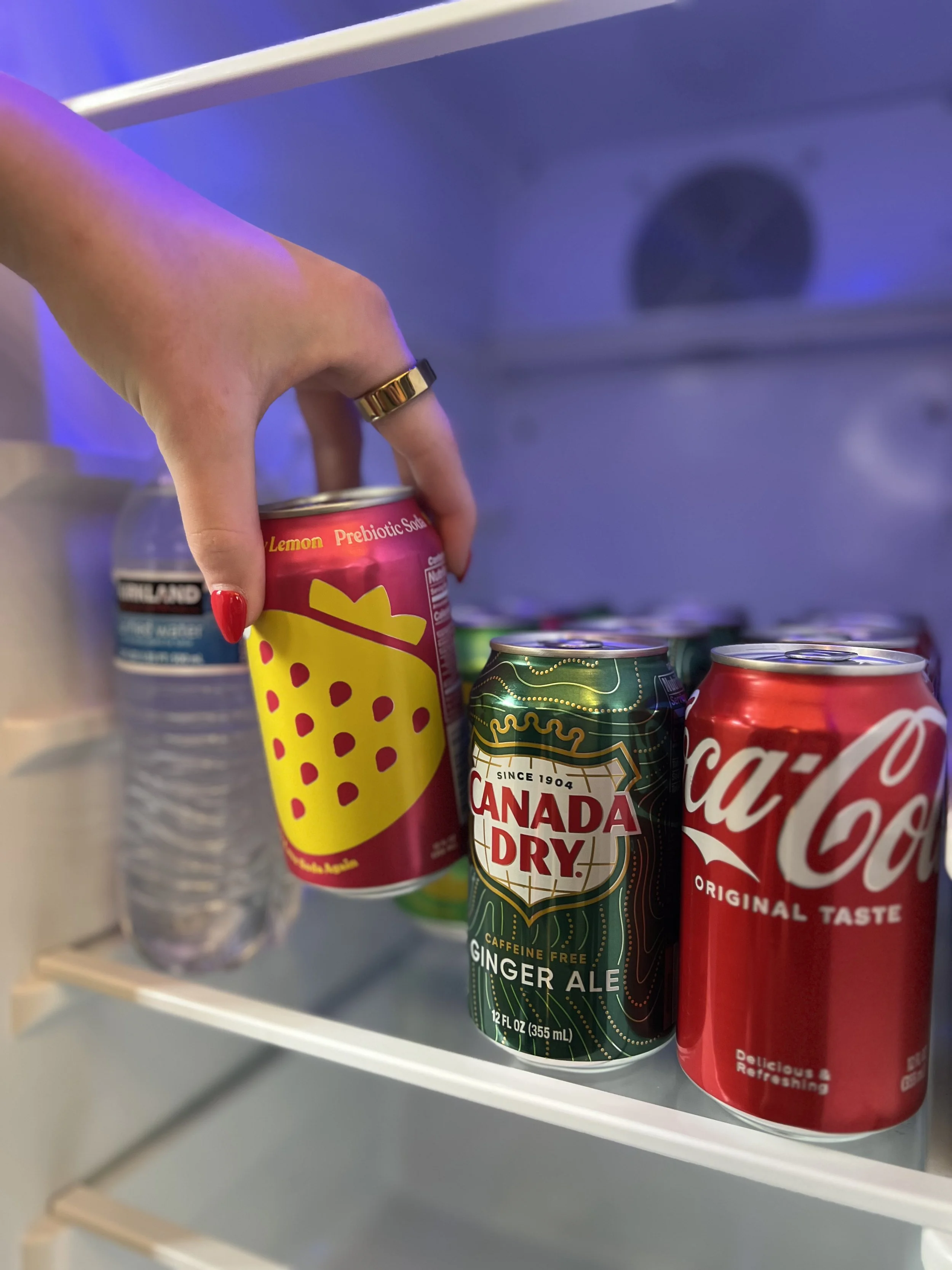 Hand reaching for a vibrant can of Olipop next to Canada Dry Ginger Ale and Coca-Cola in a refrigerator.