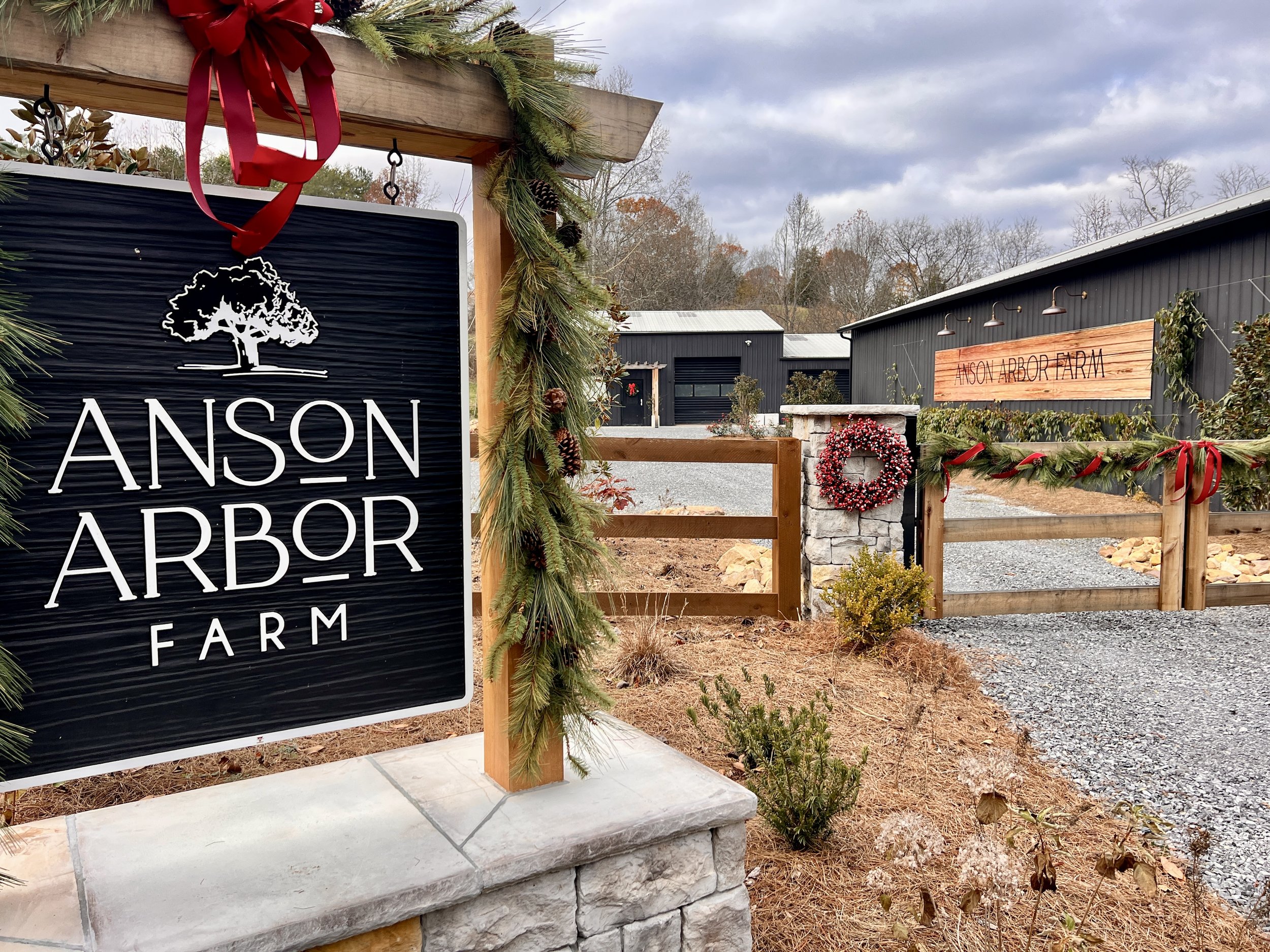 Entrance of Anson Arbor Farm decorated for the holidays, featuring a sign with a tree logo, a wreath on a stone pillar, and garlands with red bows.