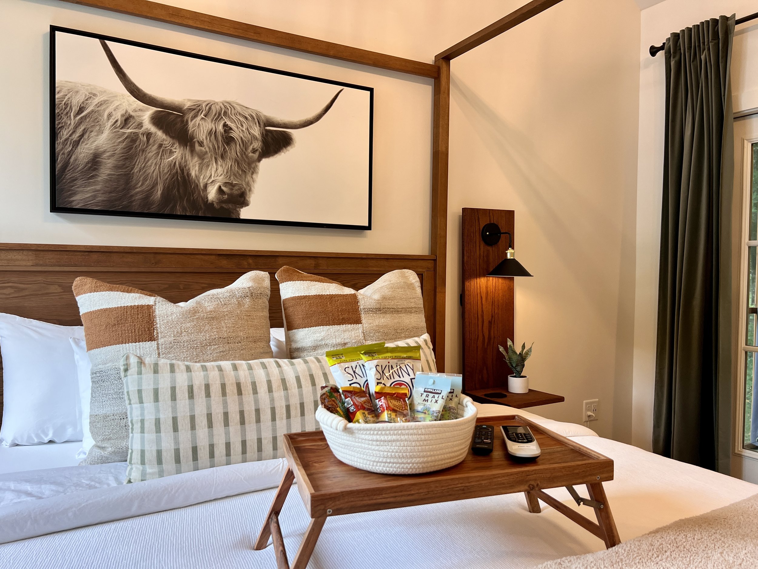 Cozy bedroom with a wooden headboard featuring a framed cow print. The bed is adorned with striped and checkered pillows. A wooden tray on the bed holds snacks, a TV remote, and a small plant. Room has warm lighting and a window with dark curtains.