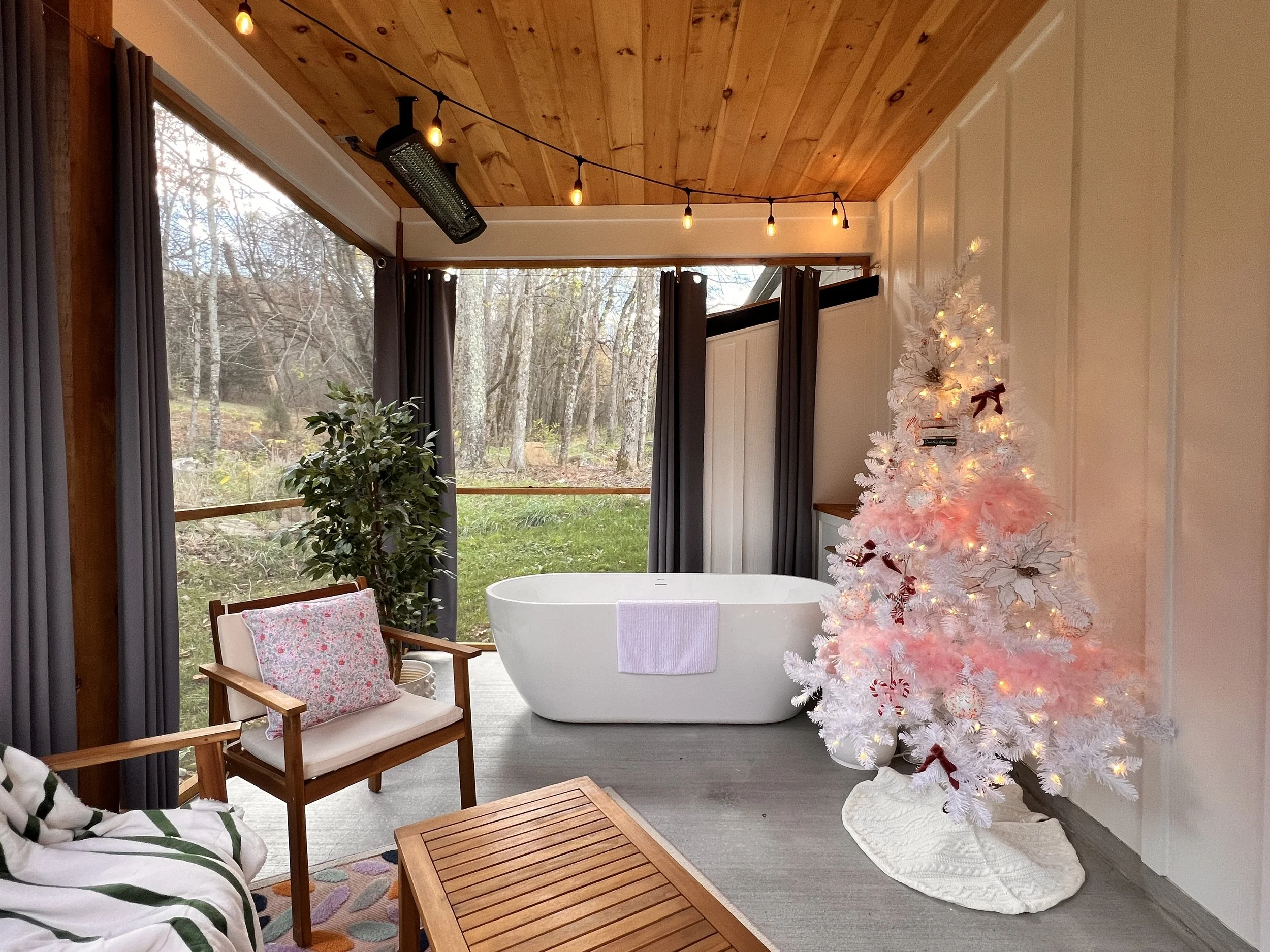 A cozy outdoor room featuring a freestanding white bathtub, a white Christmas tree with pink and white decorations, a wooden chair with a floral cushion, and a wooden table. The ceiling is made of wood with string lights hanging. Large windows reveal