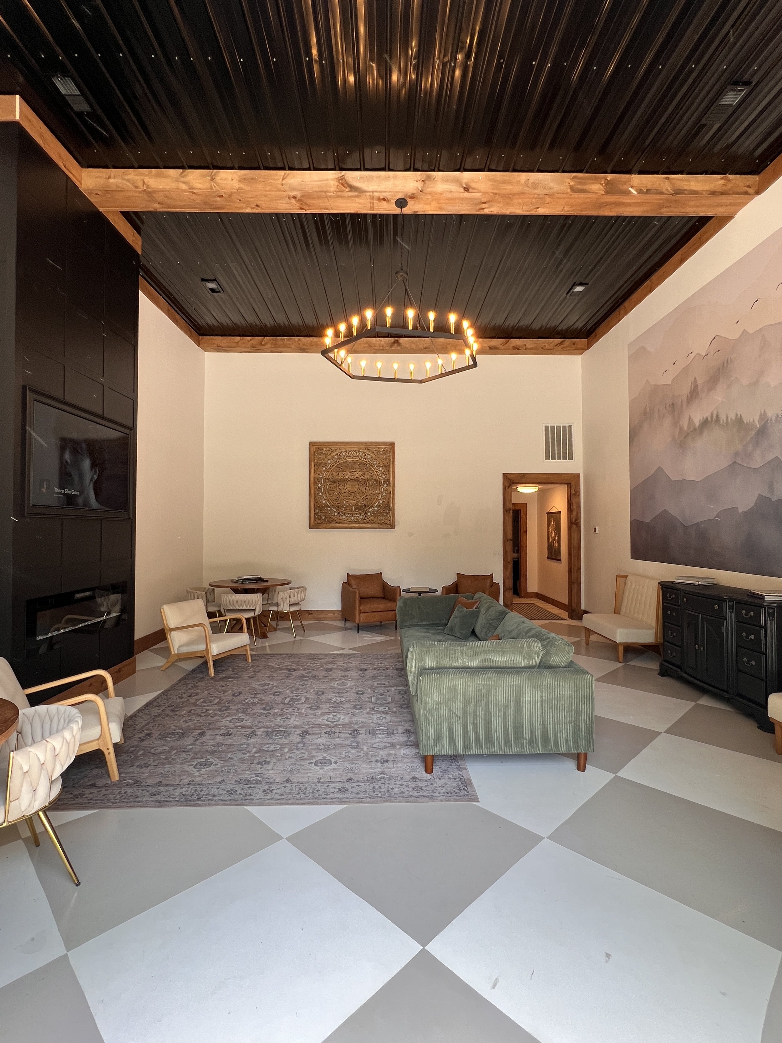 Contemporary living room with a black paneled ceiling and exposed wooden beams. Features a large chandelier, green sofa, chairs, a patterned area rug, a dining table, and wall decorations. The walls have a mountain mural and a decorative panel. The f