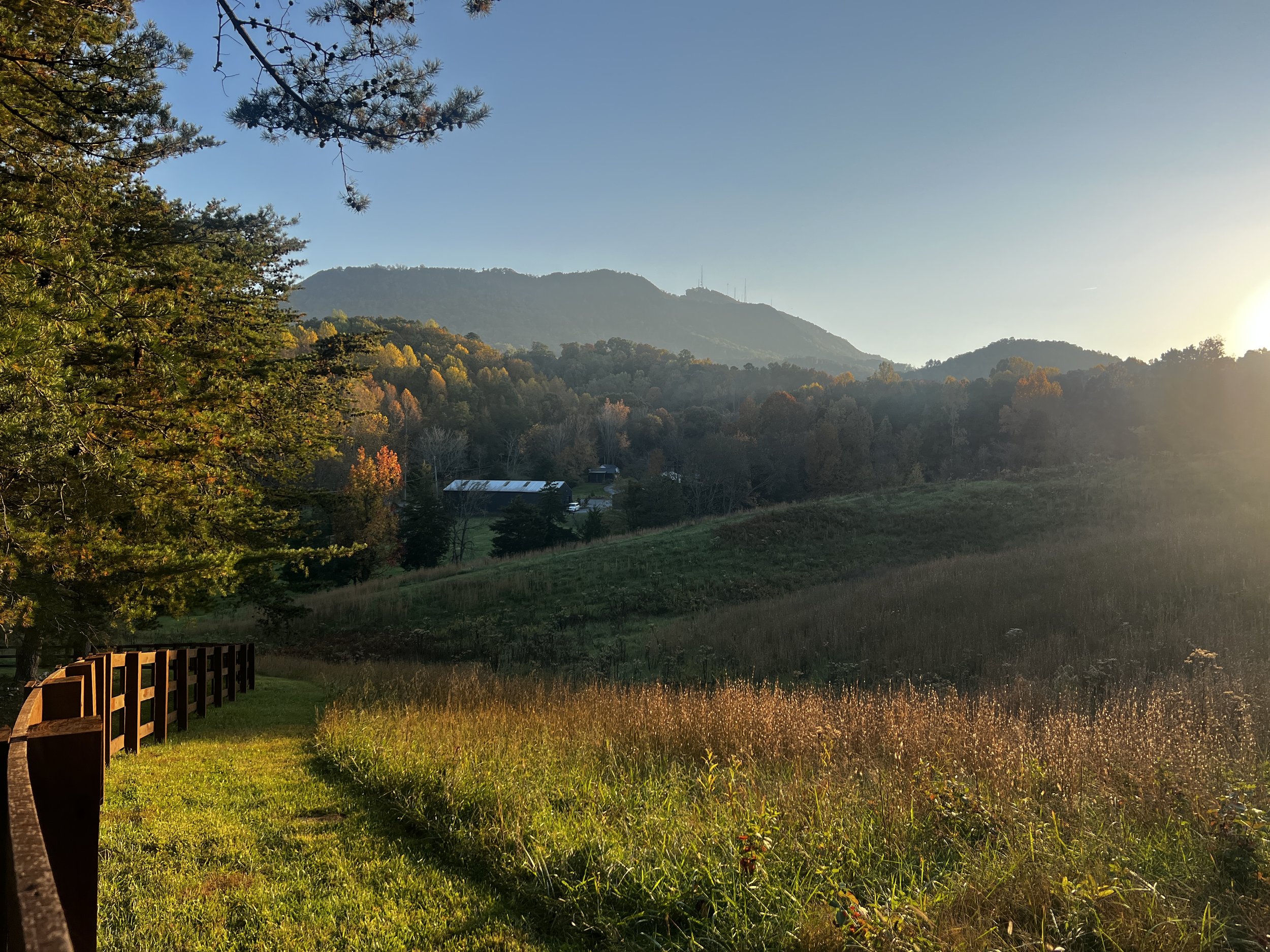 Sunlit rural landscape with a grassy field, trees, distant hills, and a small barn in the background.