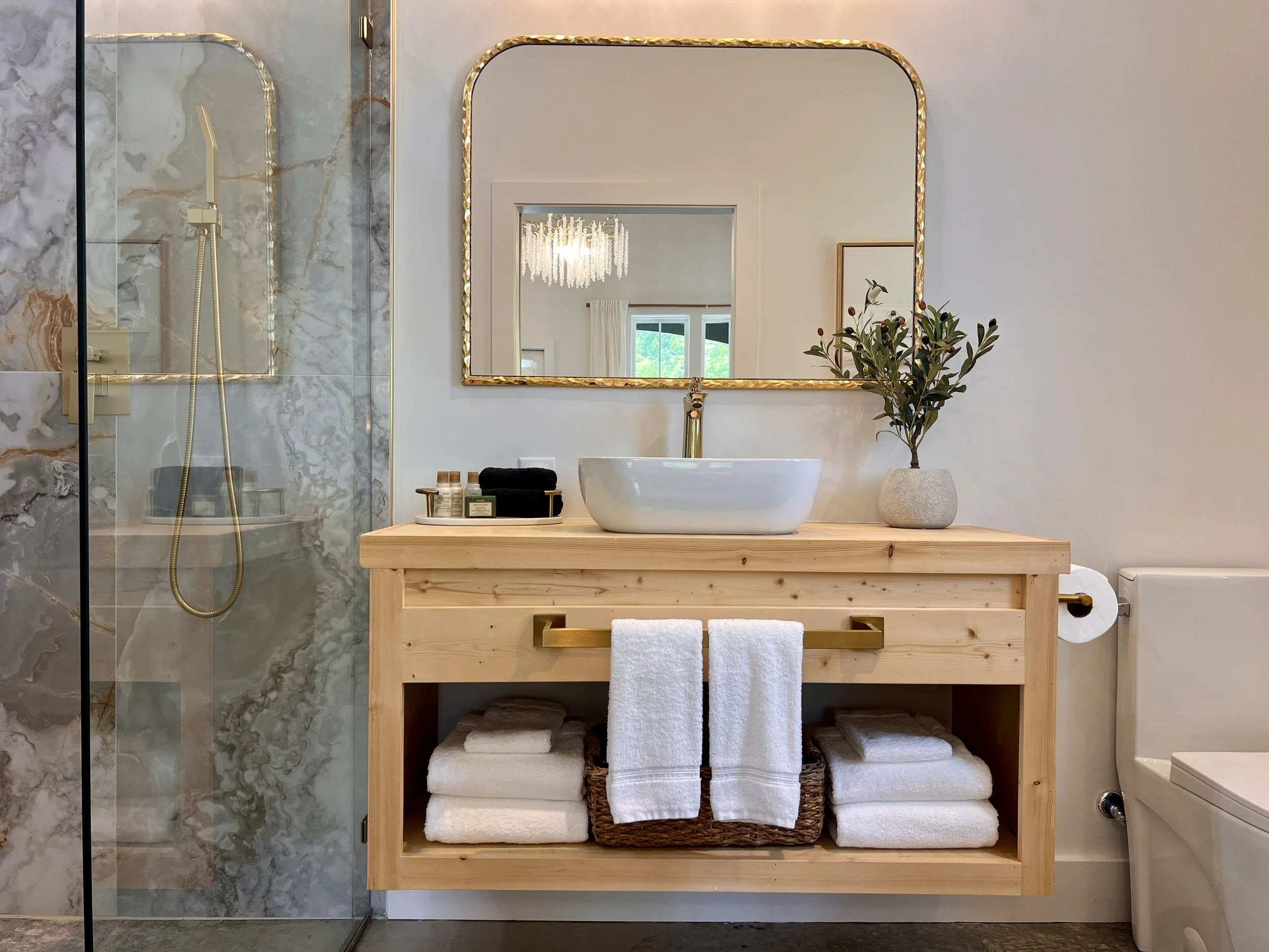 Bathroom interior with a wooden vanity, white vessel sink, and gold-framed mirror. A glass-enclosed shower with marble walls is to the left. Towels and toiletries are neatly arranged below the sink. A plant in a stone vase decorates the vanity.