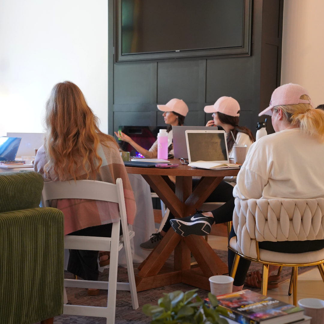 Group of women sitting around a table in a meeting room, some wearing pink caps, with laptops, notebooks, and water bottles.