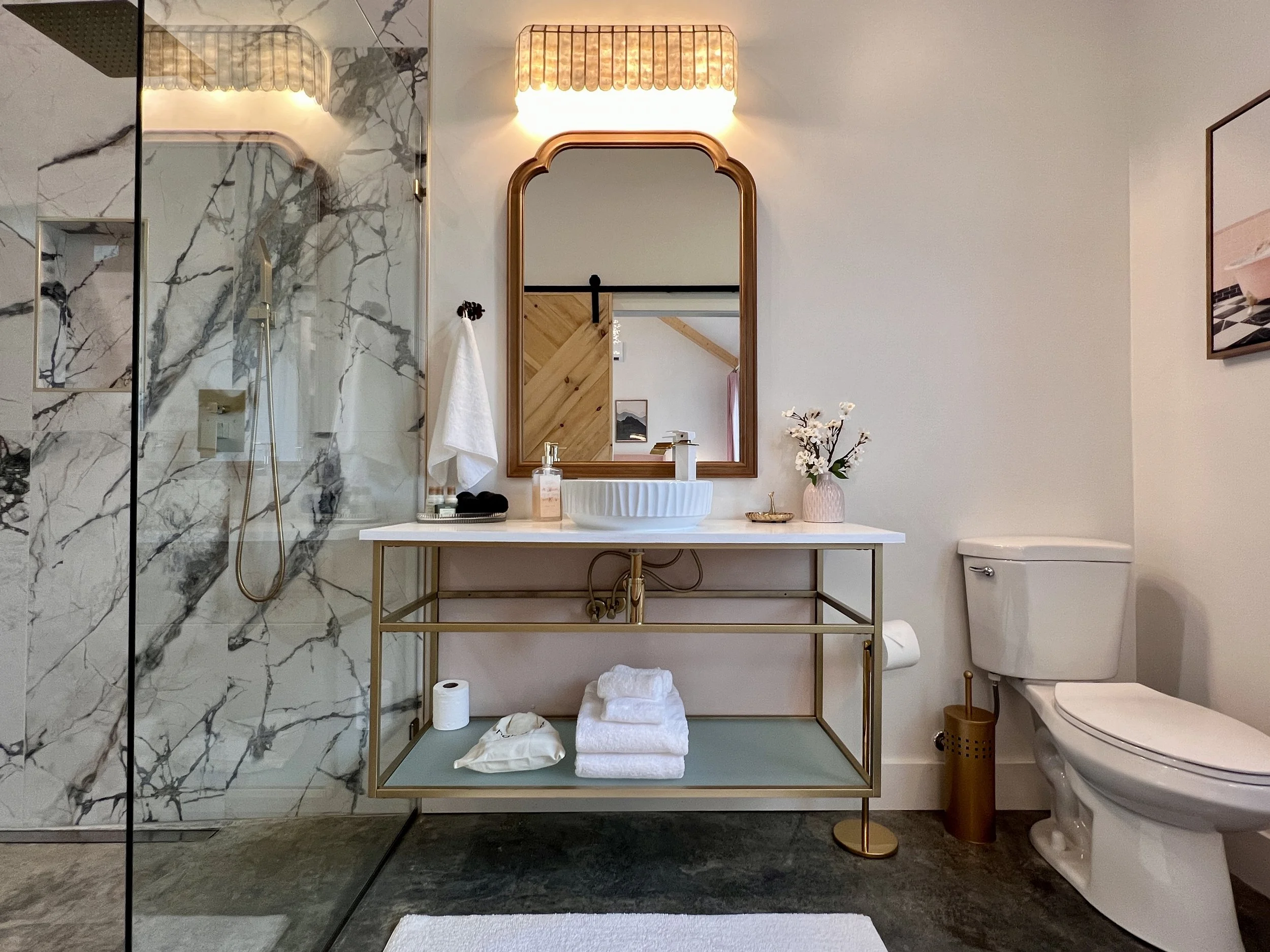 Stylish modern bathroom with a marble shower, vanity with white basin, decorative mirror, toilet, and shelf with towels. Neutral color scheme and elegant lighting.