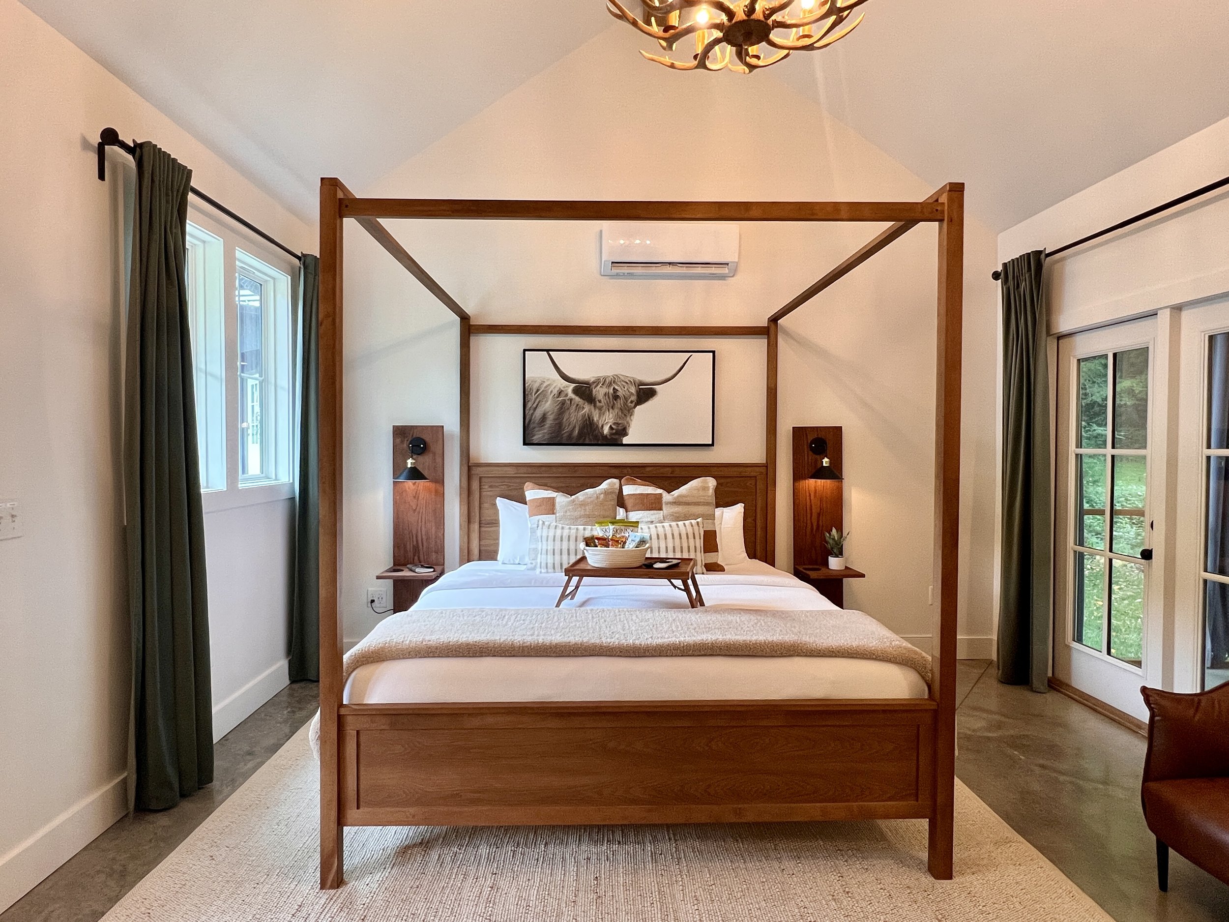 Cozy bedroom with a wooden canopy bed, neatly arranged pillows, bedside tables, a large animal photograph above the bed, green curtains, and an antler chandelier.