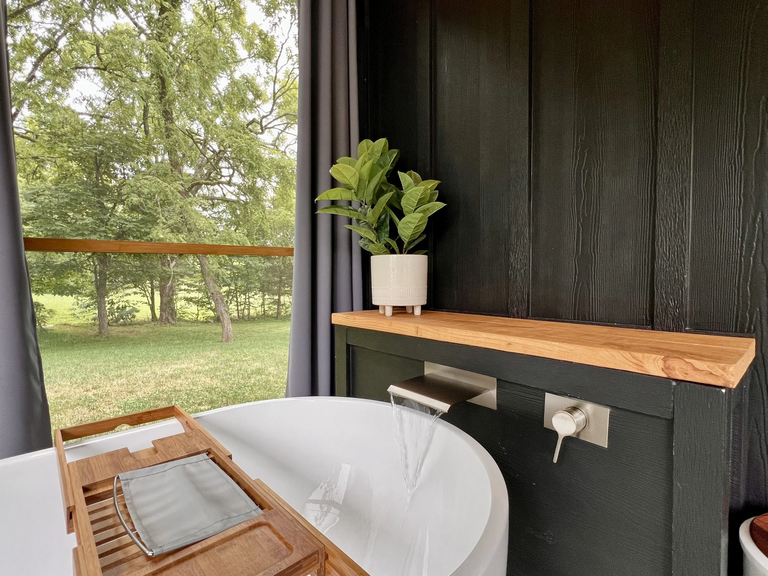 Modern bathroom interior featuring a white bathtub with a wooden tray, a sleek faucet, and a potted plant on a wooden shelf. Large window with views of green trees and grass.