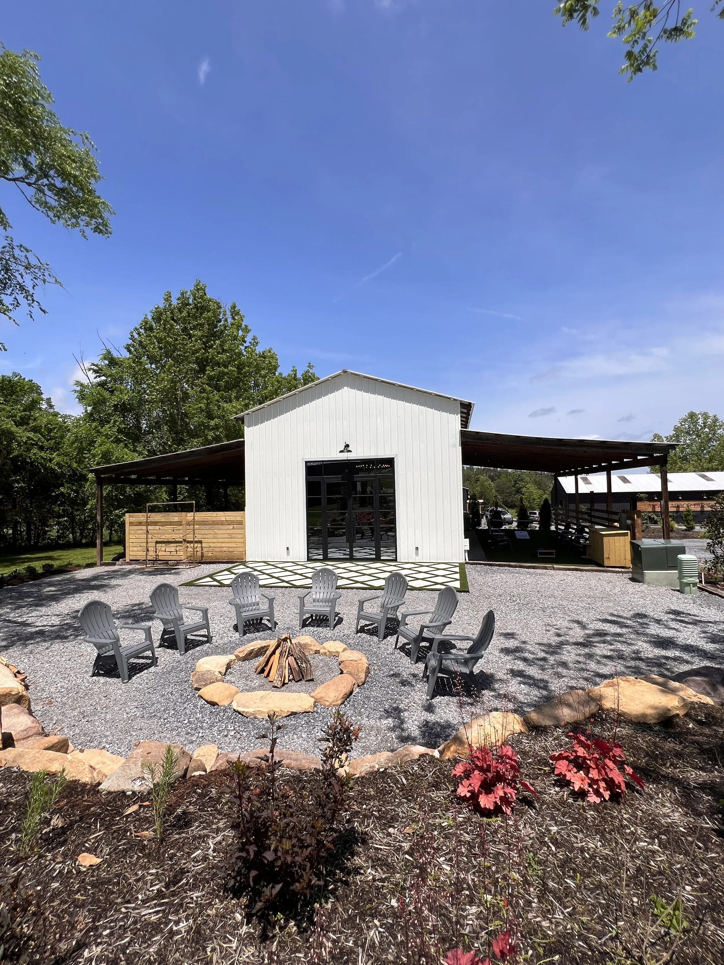 Outdoor seating area with plastic chairs arranged around a fire pit in front of a white barn-style building, surrounded by gravel and landscaping, with trees in the background under a clear blue sky.