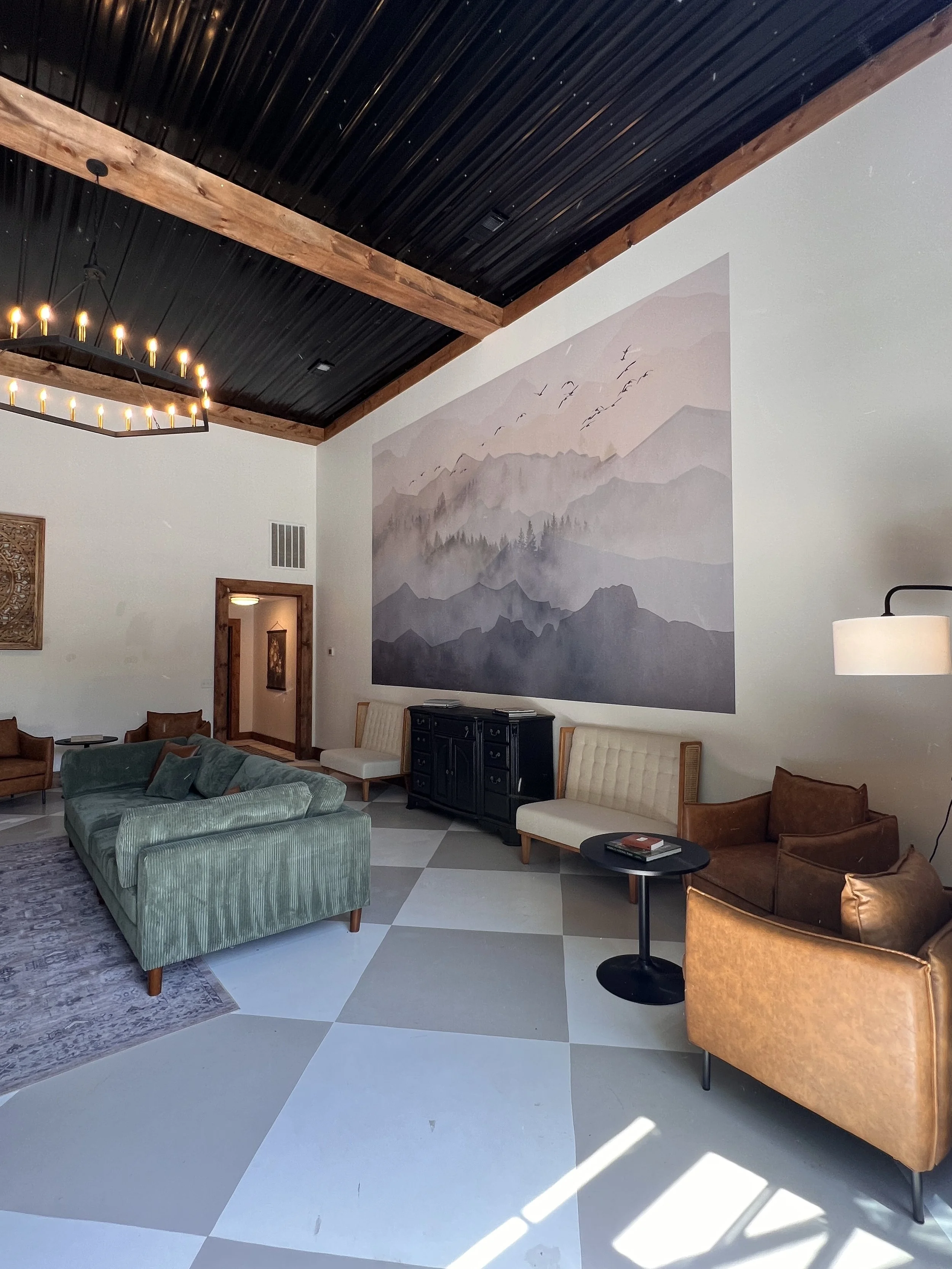 Modern lounge area with green sofa, brown leather chairs, large wall mural of mountains, and black ceiling with wooden beams.