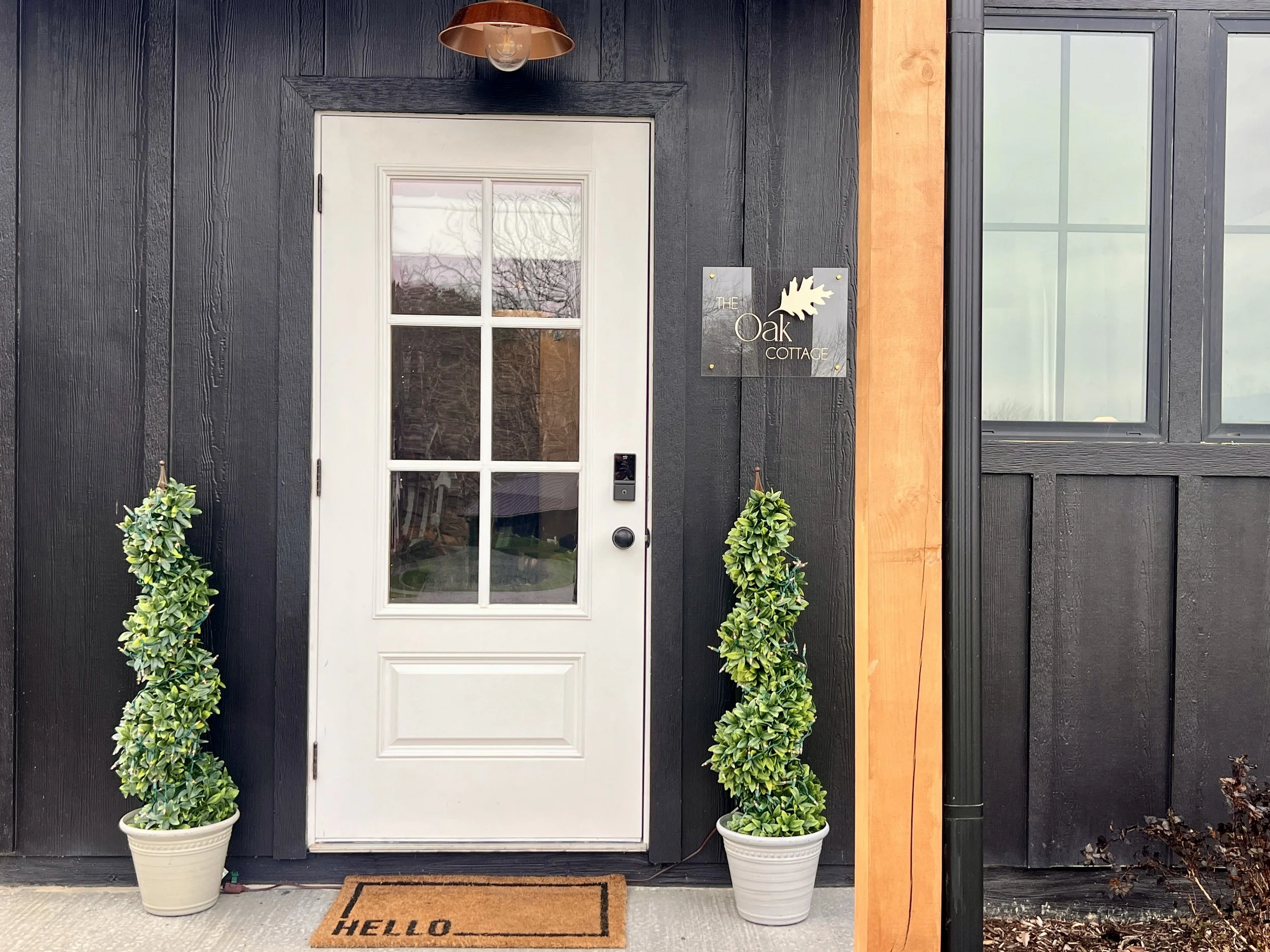Front door with large glass panels, flanked by two potted topiary plants, a "Hello" welcome mat, and a sign reading "The Oak Cottage."
