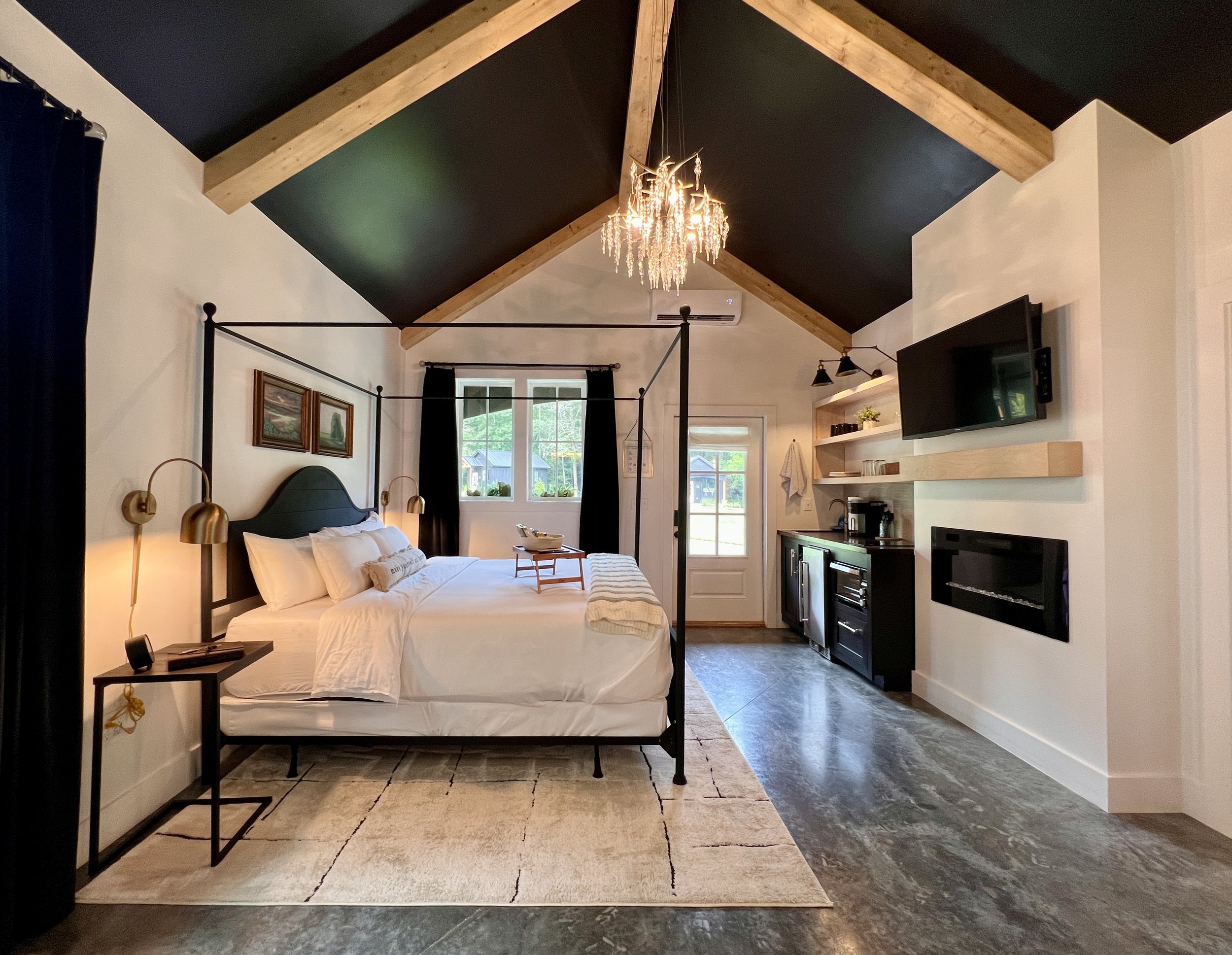 Modern bedroom with a vaulted ceiling, featuring a four-poster bed, chandelier, wall-mounted TV, and a kitchenette on the side.