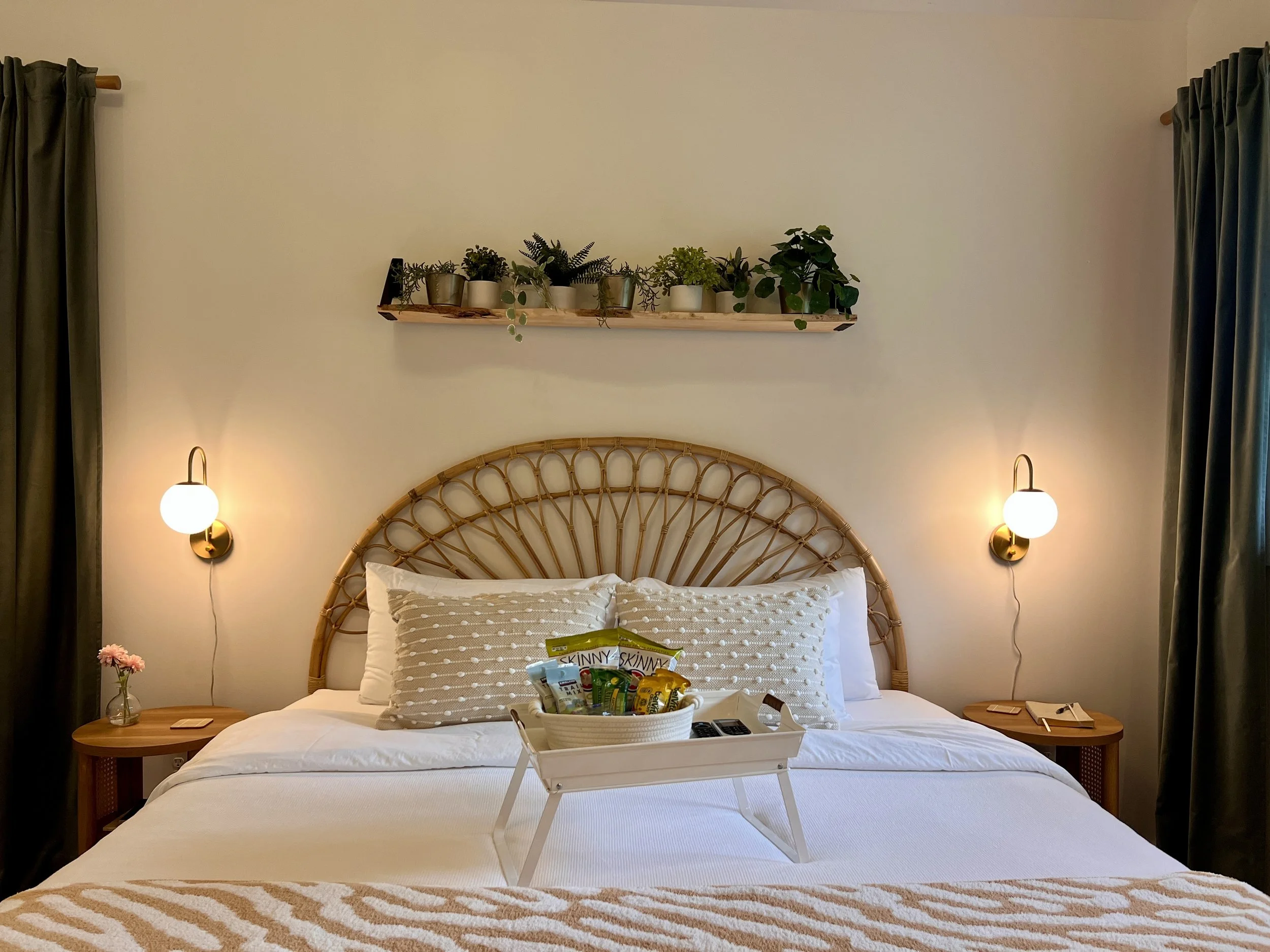 Bedroom interior with a rattan headboard, white bedding, and a decorative pillow. A white tray with snacks and a plant is on the bed. Wall-mounted lights are above two wooden nightstands with a vase and a notepad. Above the bed, a shelf with potted p