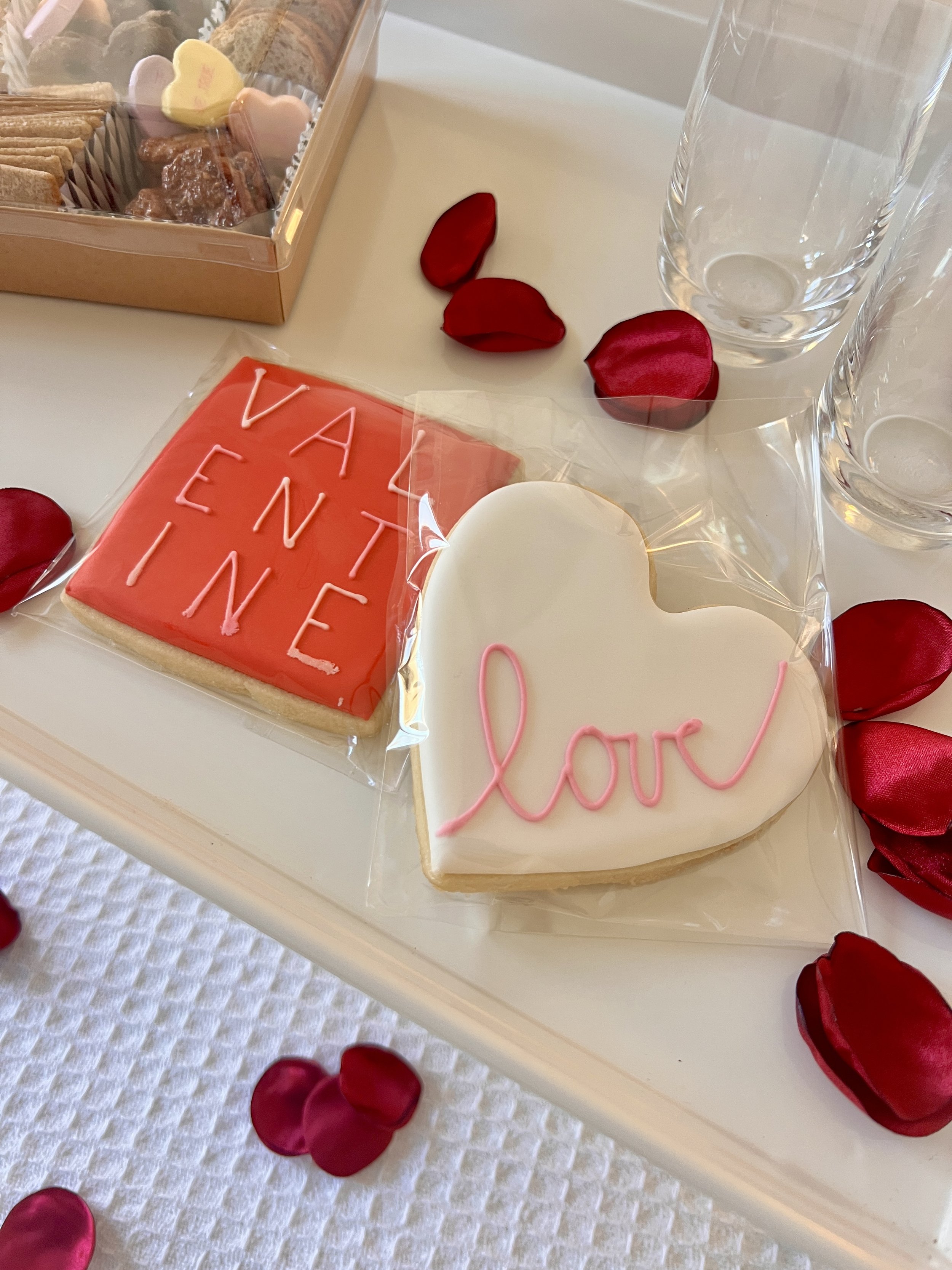 Valentine's Day themed cookies on a tray with red rose petals and glass cups.
