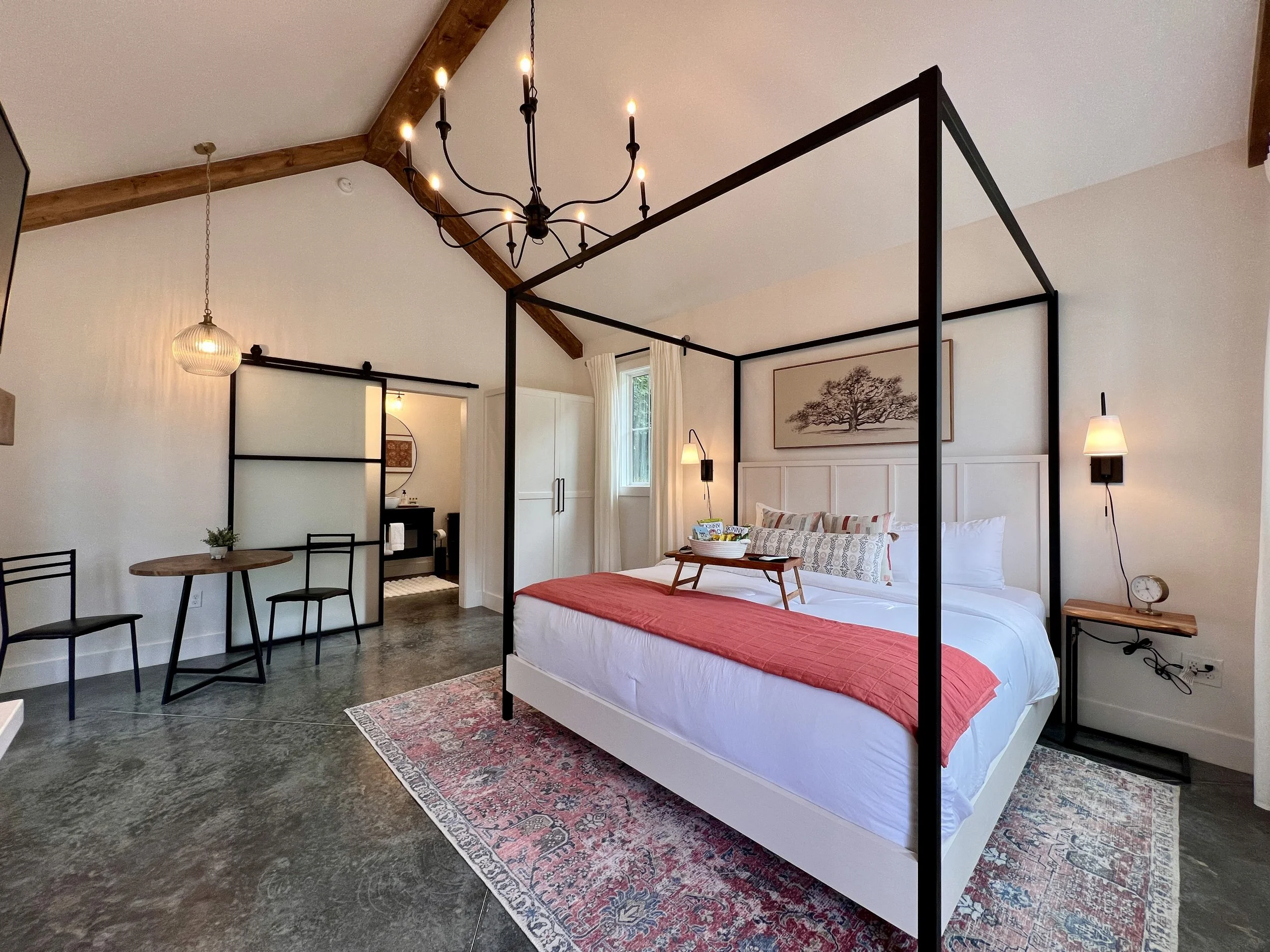 Modern bedroom with a four-poster bed, decorated with white and red bedding and decorative pillows. Room includes a rustic chandelier, a small dining table with two chairs, and a sliding barn door leading to an ensuite bathroom. The room features a m