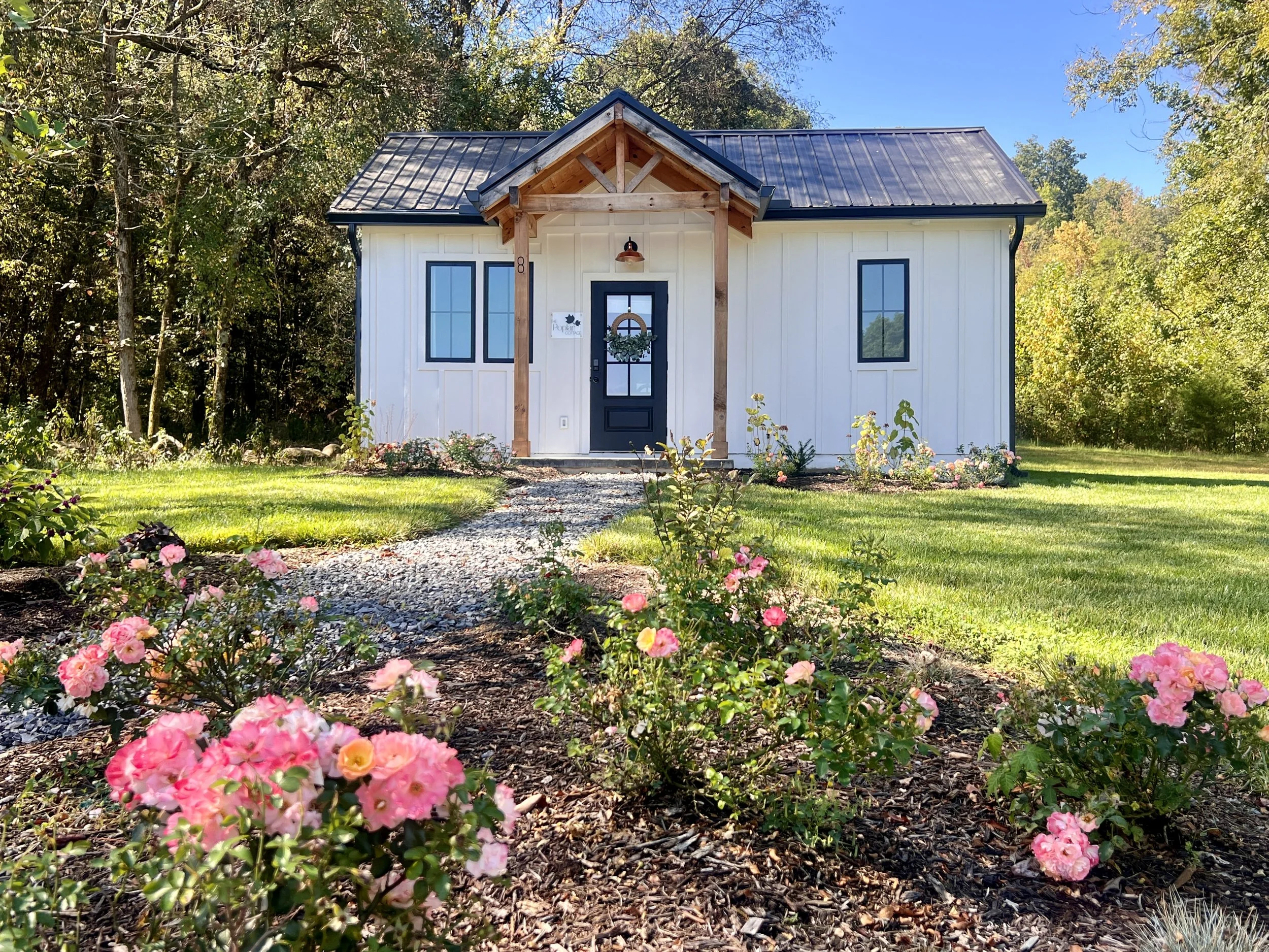 Small white cabin with a metal roof surrounded by a garden with pink roses and green lawn, set in a wooded area.