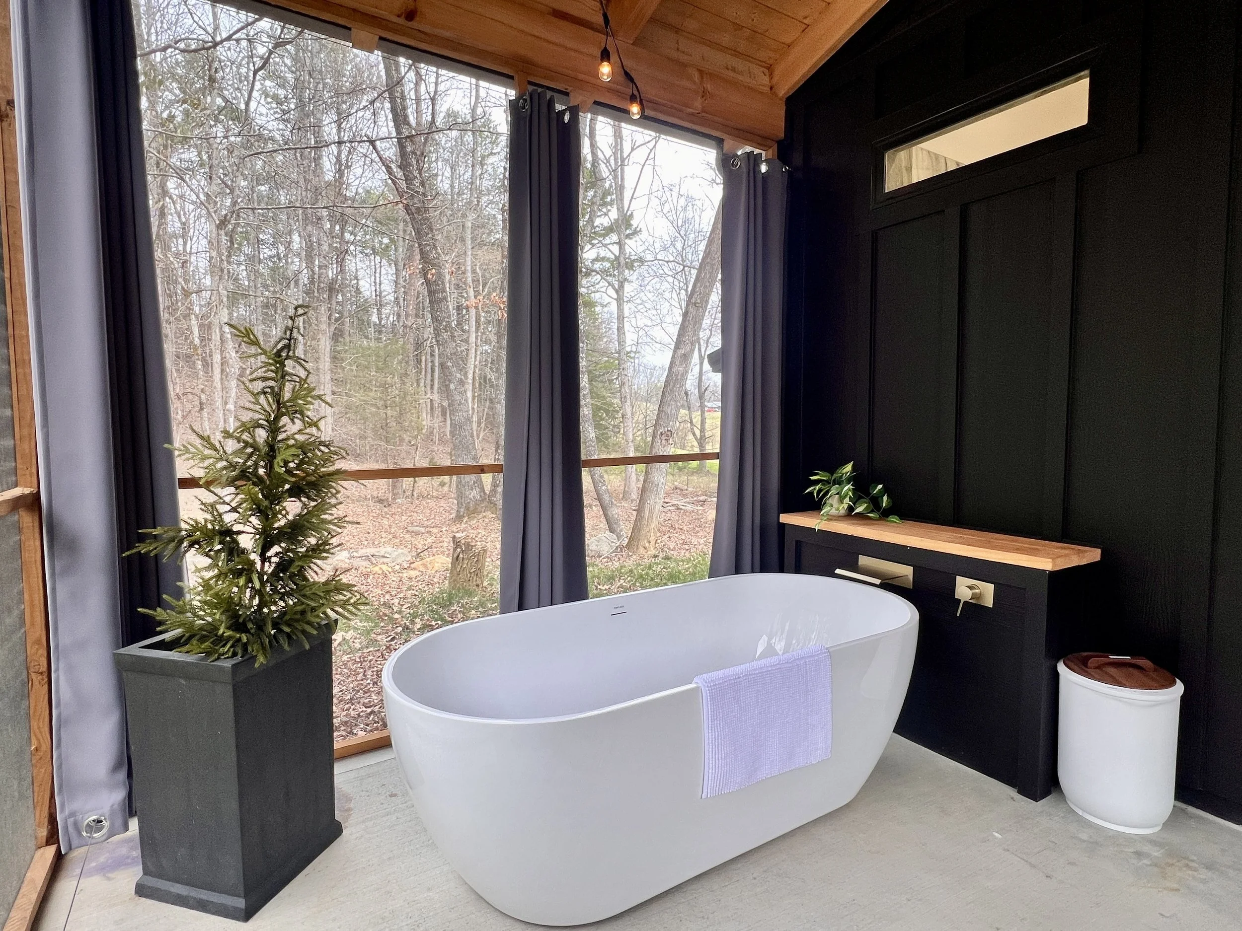 Outdoor bathroom with a modern white freestanding bathtub, dark curtains, wooden ceiling, a potted plant, and a wooden bench, overlooking a wooded area.