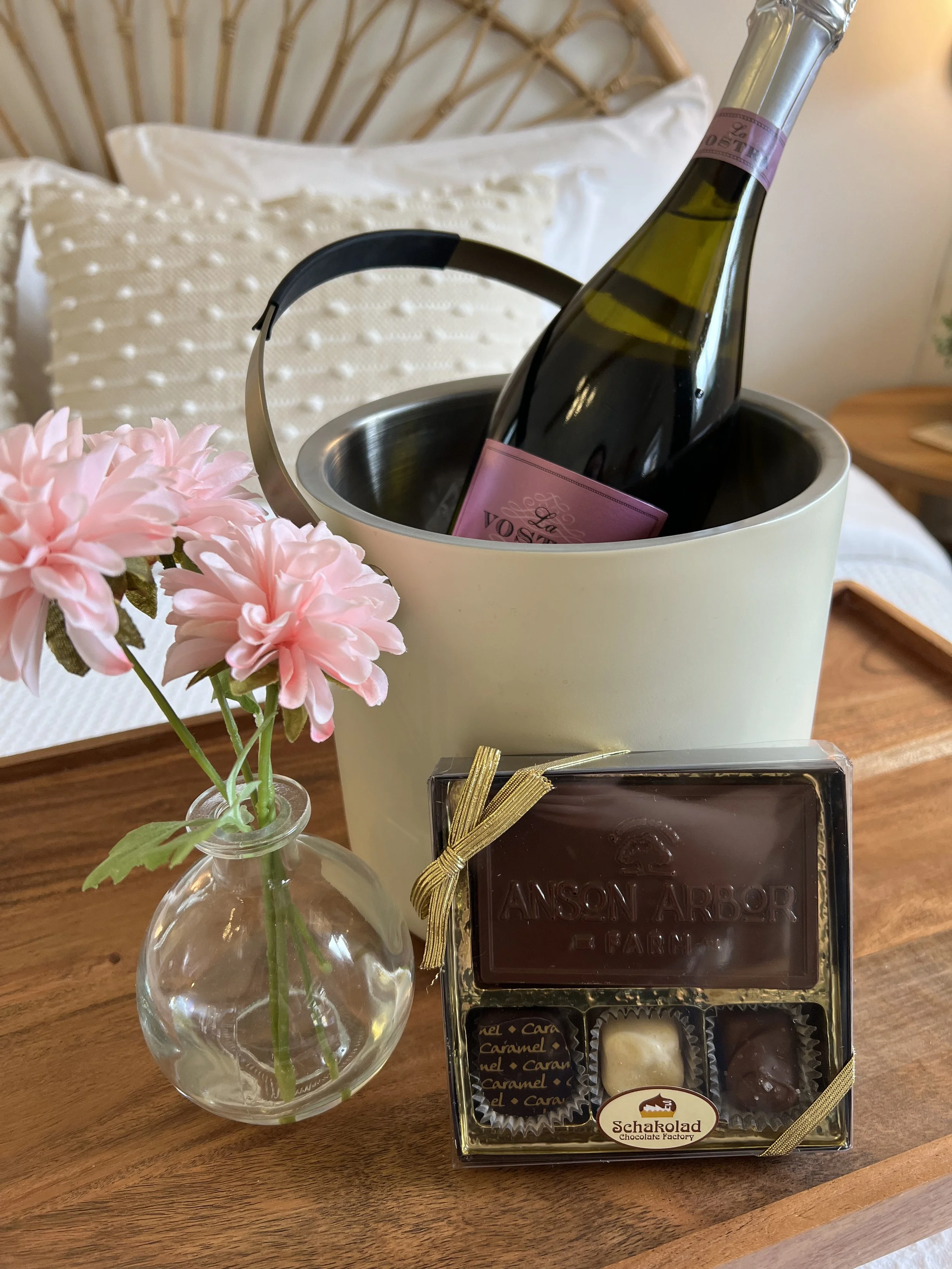 Bottle of sparkling wine in an ice bucket, a box of chocolates with "Anson Arbor" label, pink flowers in a vase on a wooden tray.