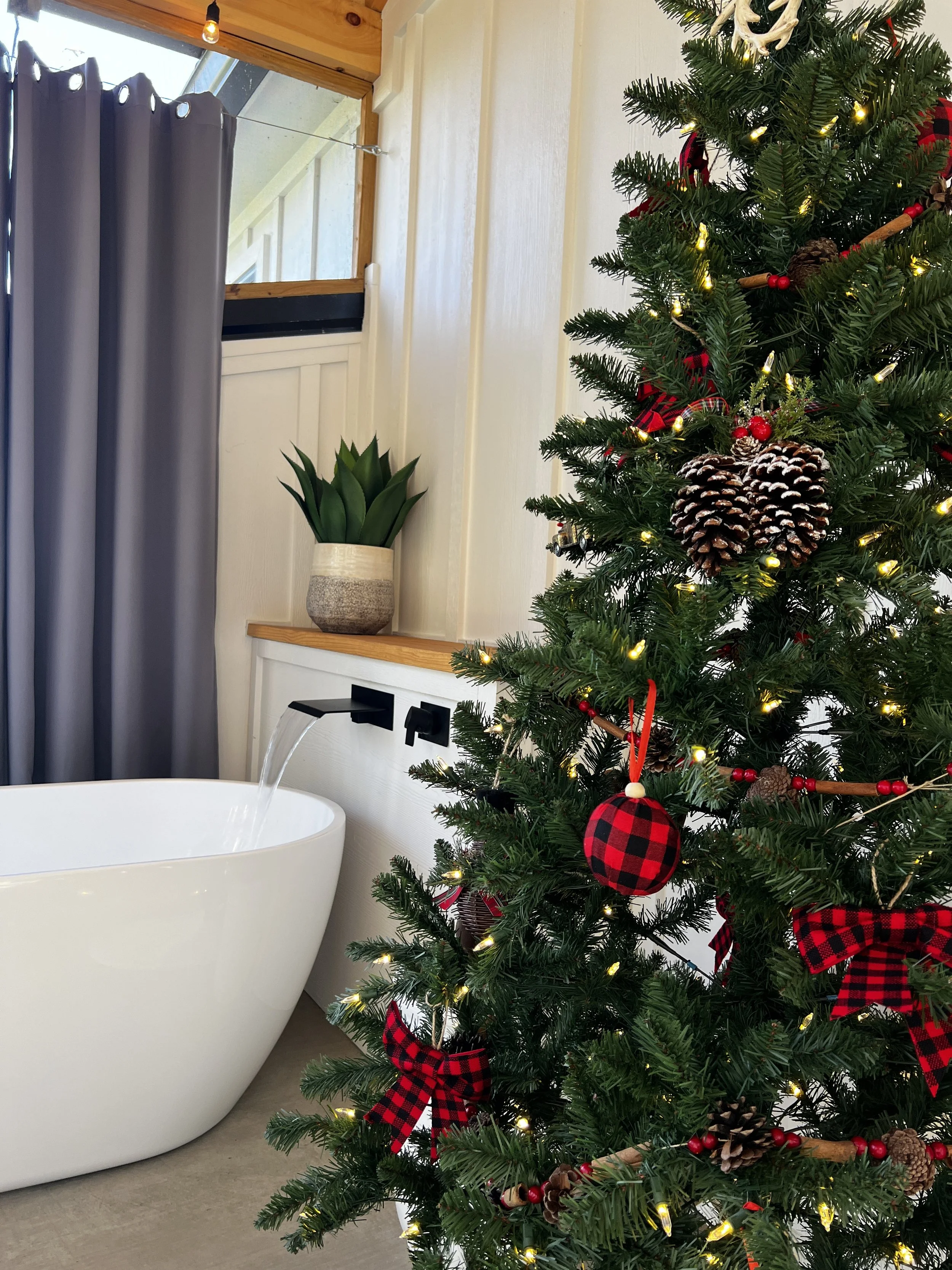A bathroom with a freestanding white bathtub and a Christmas tree decorated with red plaid ornaments and pinecones. The room has gray curtains and a small potted plant on a wooden shelf next to the tub.