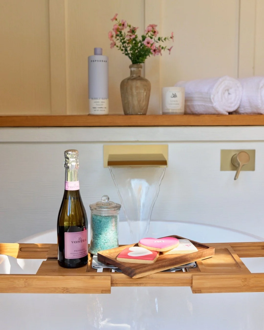 Luxurious bathroom with a wooden bathtub tray holding a bottle of sparkling wine, jar of bath salts, and heart-shaped cookies. A waterfall faucet fills the tub. The background has neatly arranged towels, a candle, flowers in a vase, and a bottle of b
