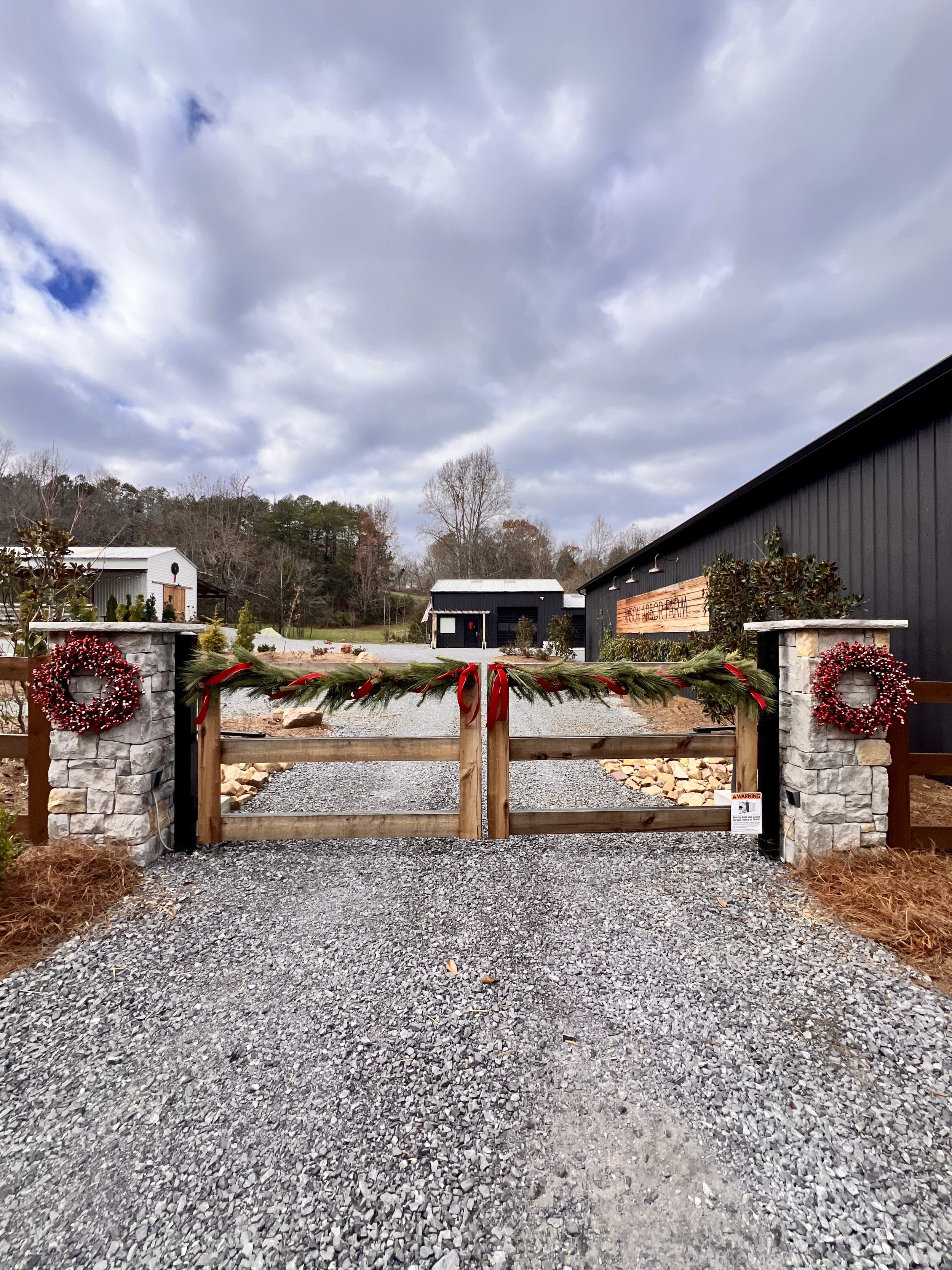 Decorative wooden gate with holiday wreaths and garland, leading to buildings against a cloudy sky.