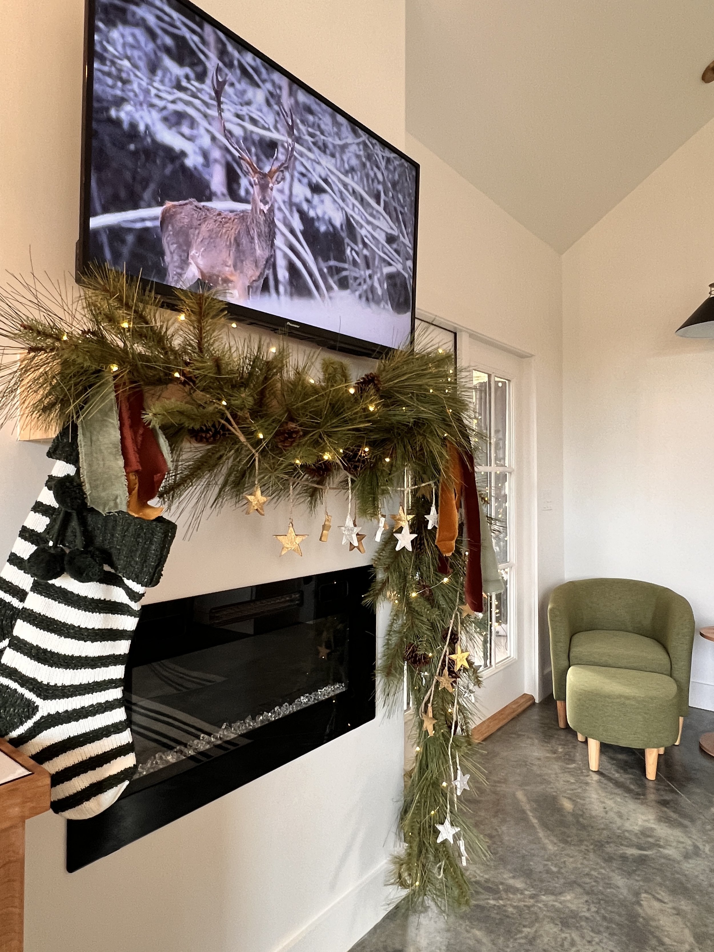 Holiday-decorated living room with a TV above a fireplace showing a deer in snow. Greenery and star decorations hang over the mantle with a striped stocking.