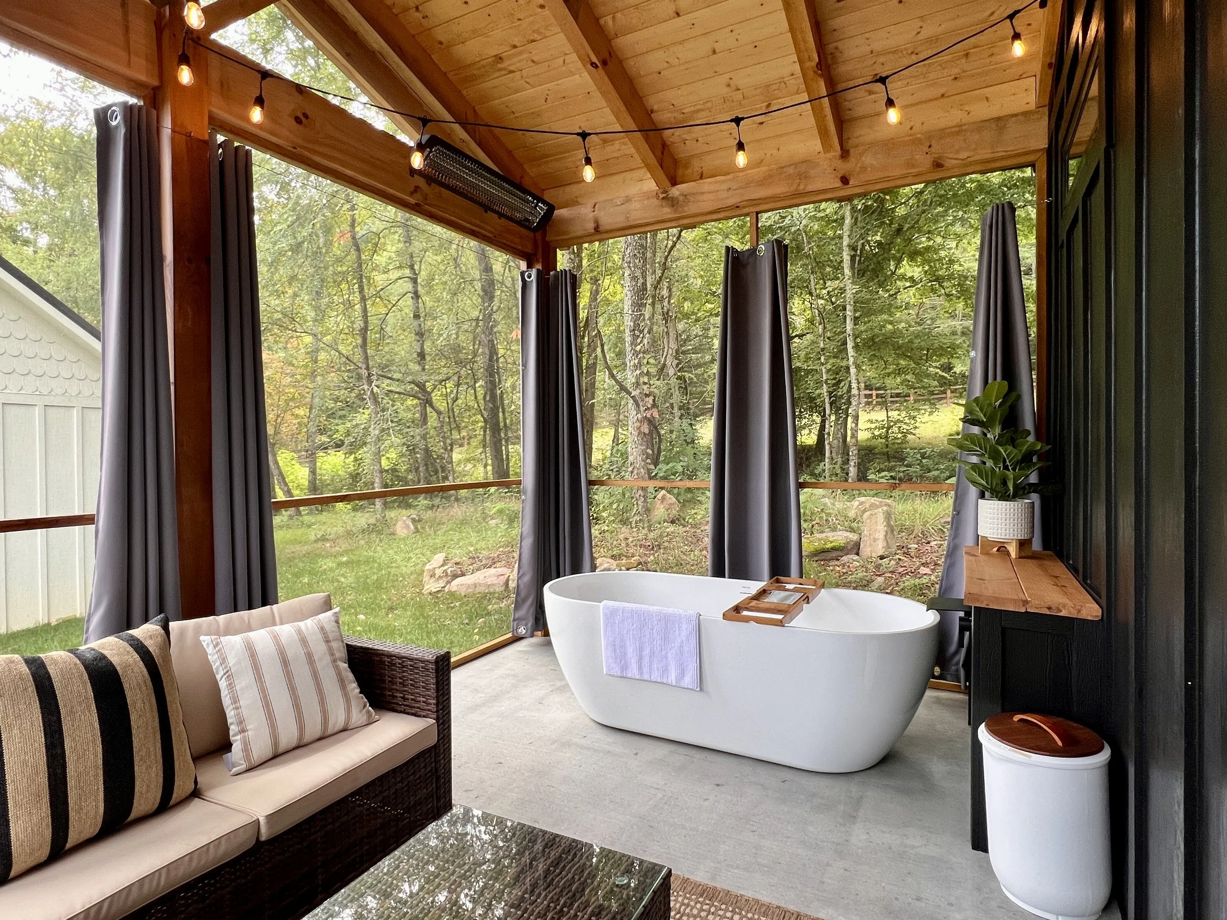 Outdoor bathroom with a freestanding bathtub, wicker sofa with cushions, string lights, and a wooden shelf with a potted plant. Surrounded by greenery.