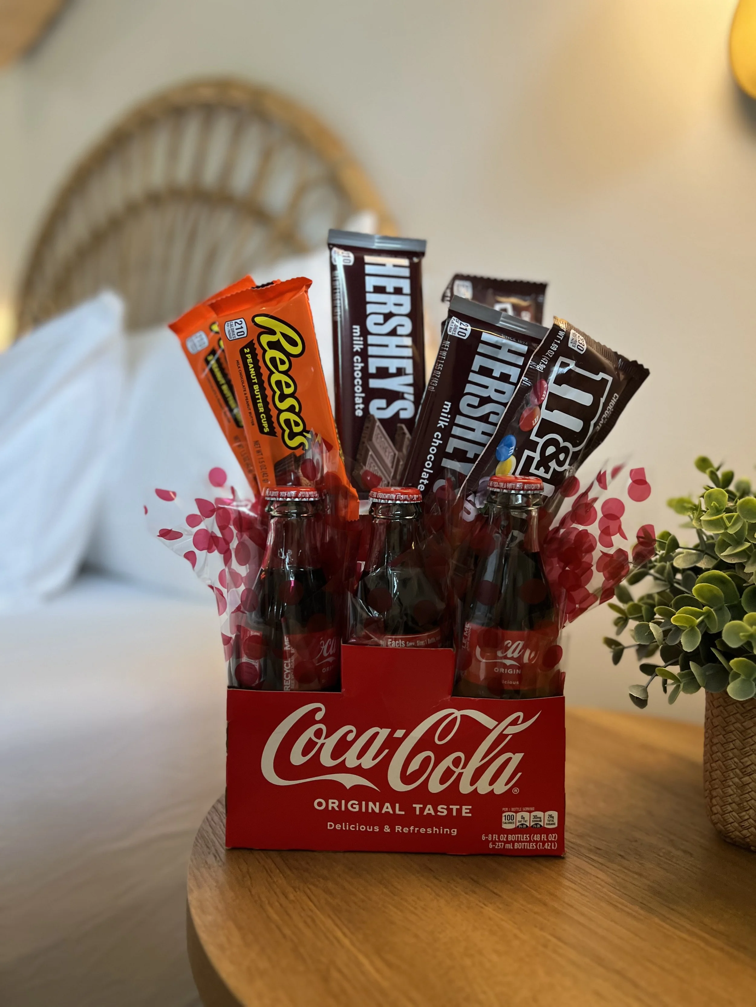 A Coca-Cola six-pack with cola bottles and various chocolate bars inserted, including Reese's, Hershey's, and M&M's, arranged on a wooden table next to a plant.