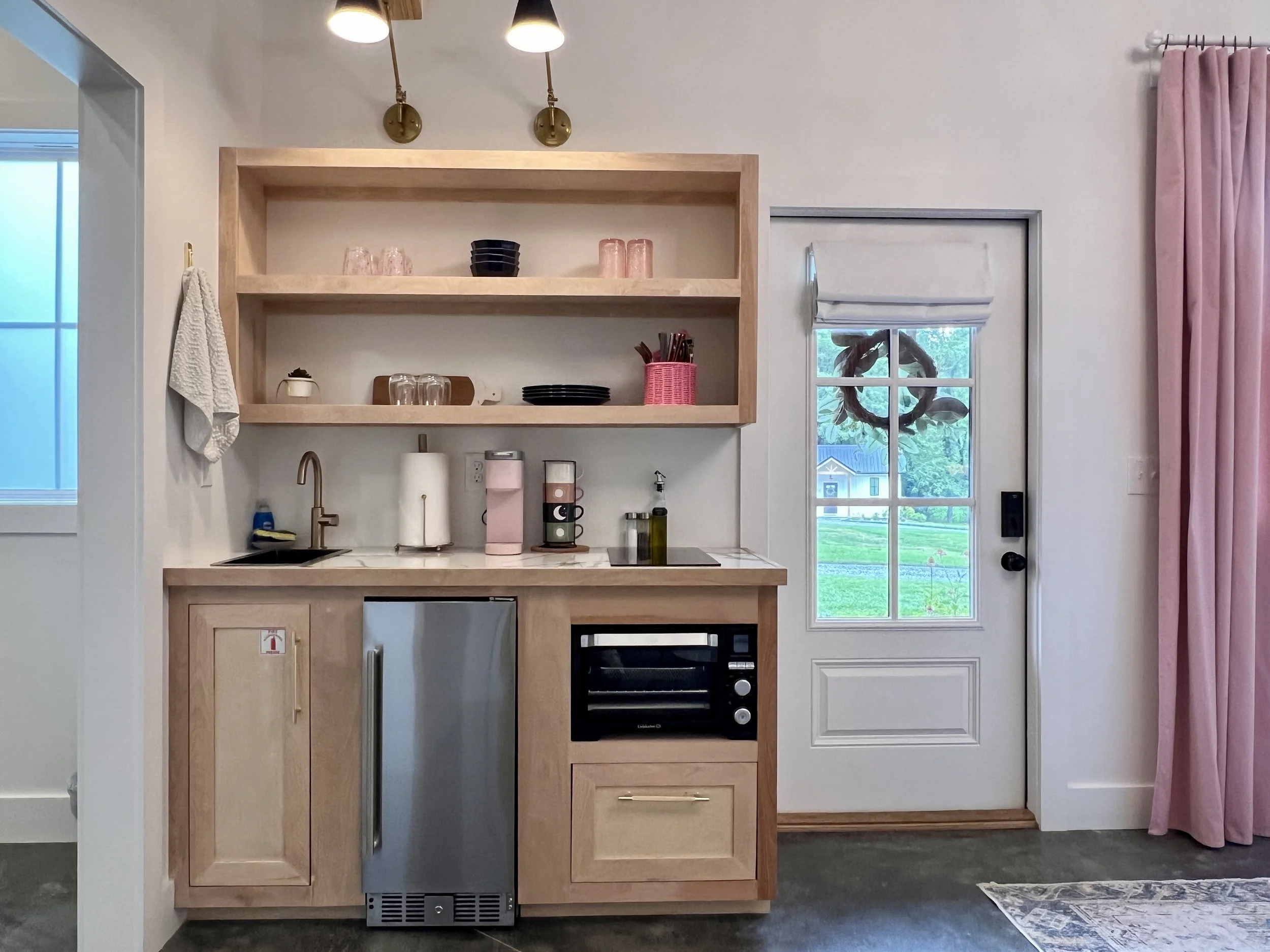 Small kitchenette with light wood cabinetry, open shelves, coffee maker, small fridge, sink, microwave, and glass door leading outside. Pink curtains frame the door.