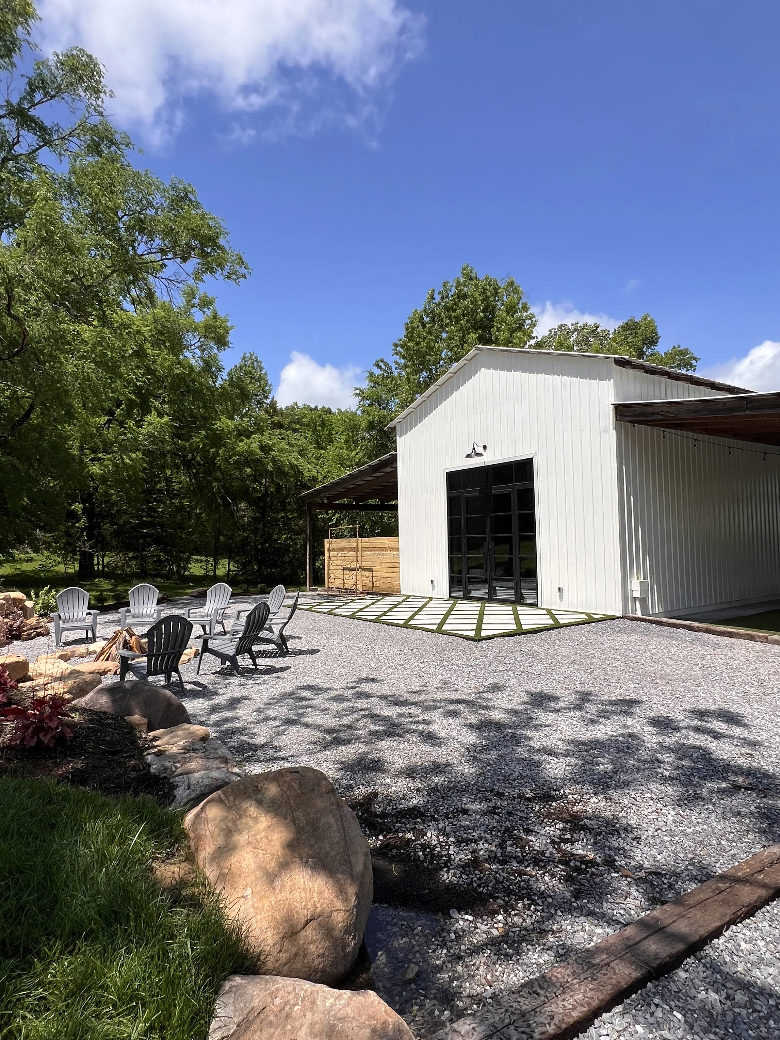 Outdoor space with a white building, gravel patio, Adirondack chairs around a fire pit, and surrounding trees under a blue sky.