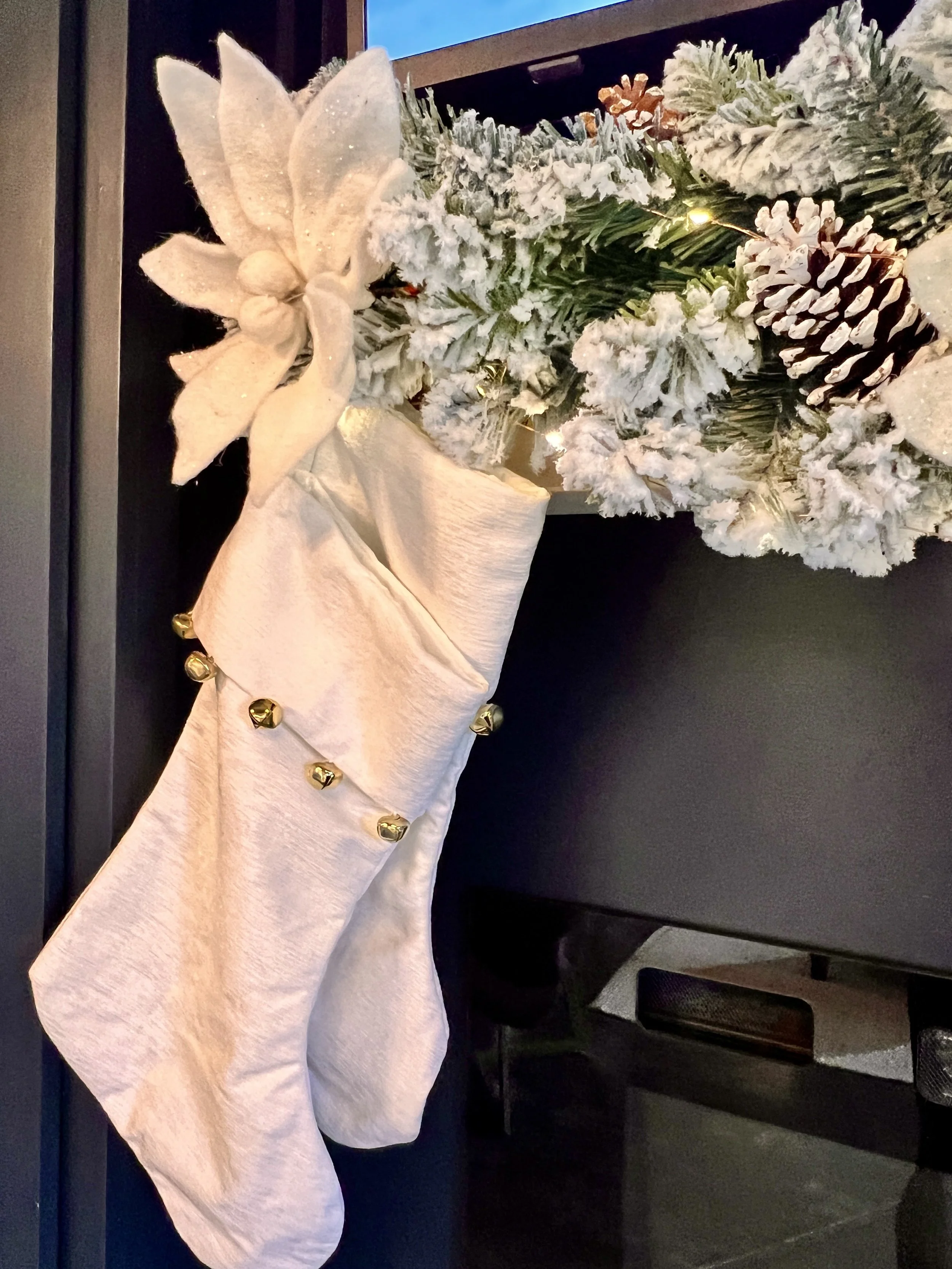 White Christmas stocking with bells hanging on a snowy wreath with pinecones and a large fabric flower.