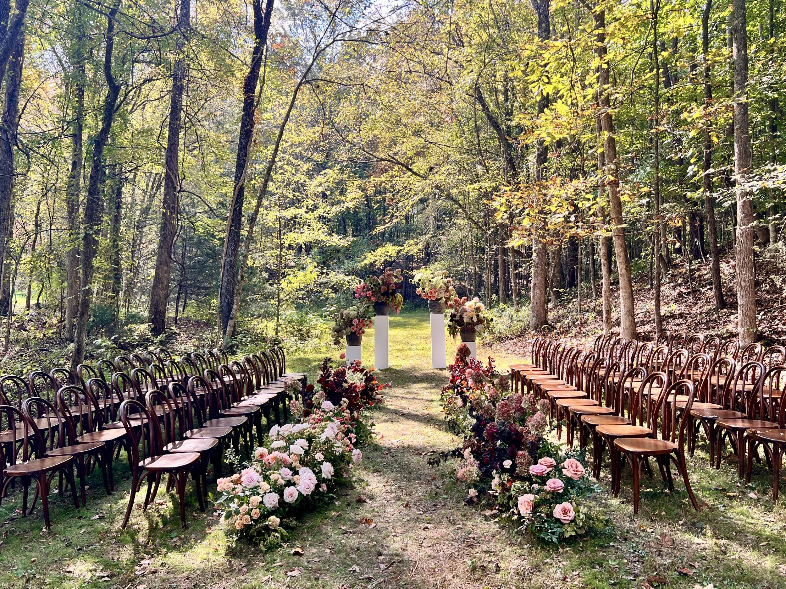 An outdoor wedding ceremony setup in a wooded area with flower arrangements and chairs on either side of a grassy aisle.