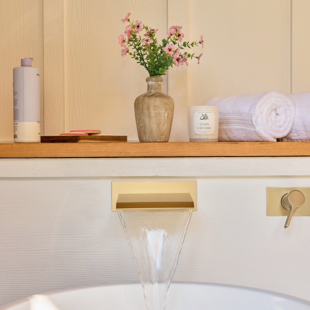 Bathroom setting with a wooden shelf featuring a vase of pink flowers, a candle labeled 'Lemon Lavender,' rolled white towels, and bath products. Below, a modern waterfall faucet fills a bathtub.