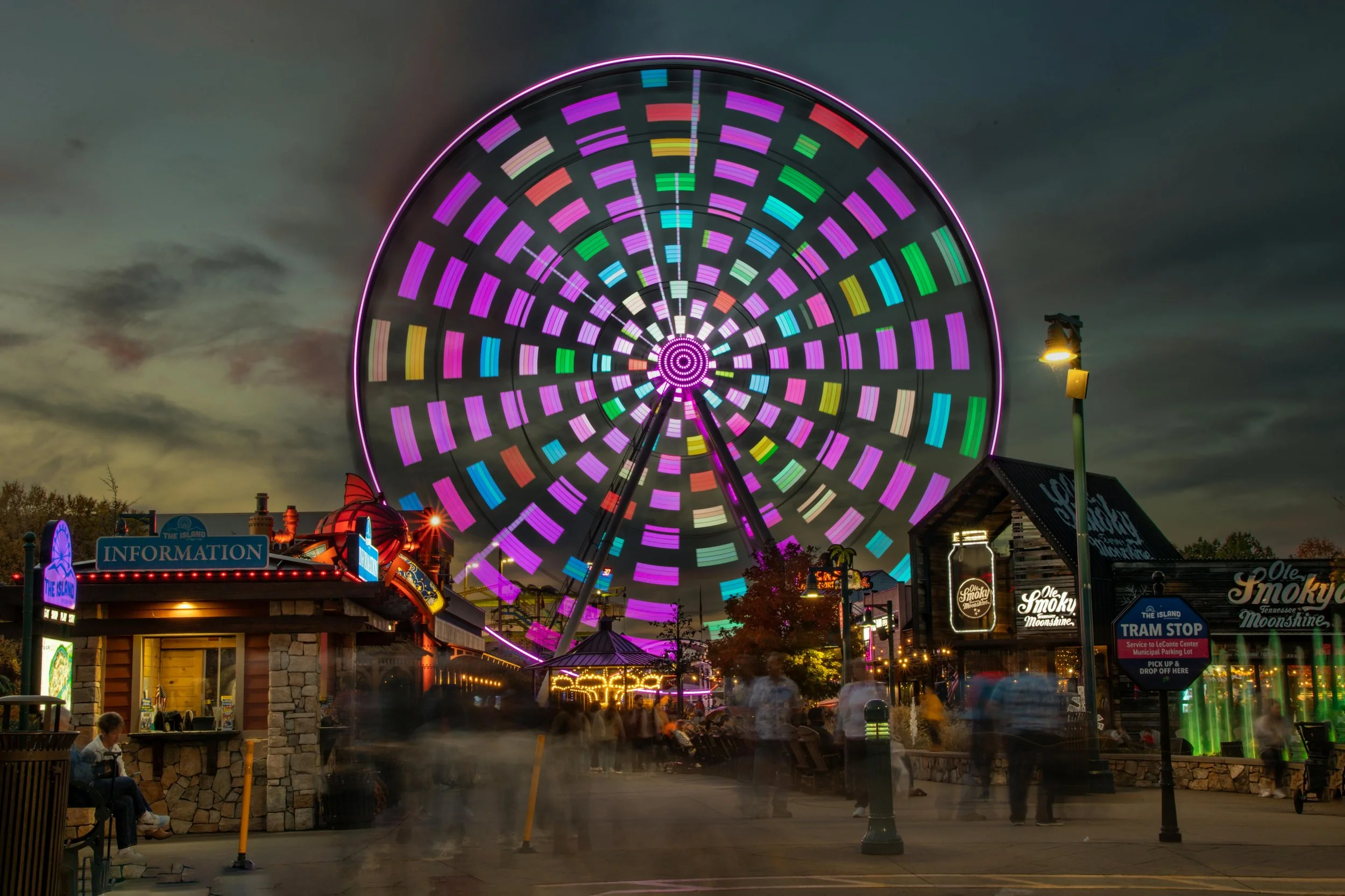 A colorful illuminated Ferris wheel at night with motion blur of people walking around in an amusement park.