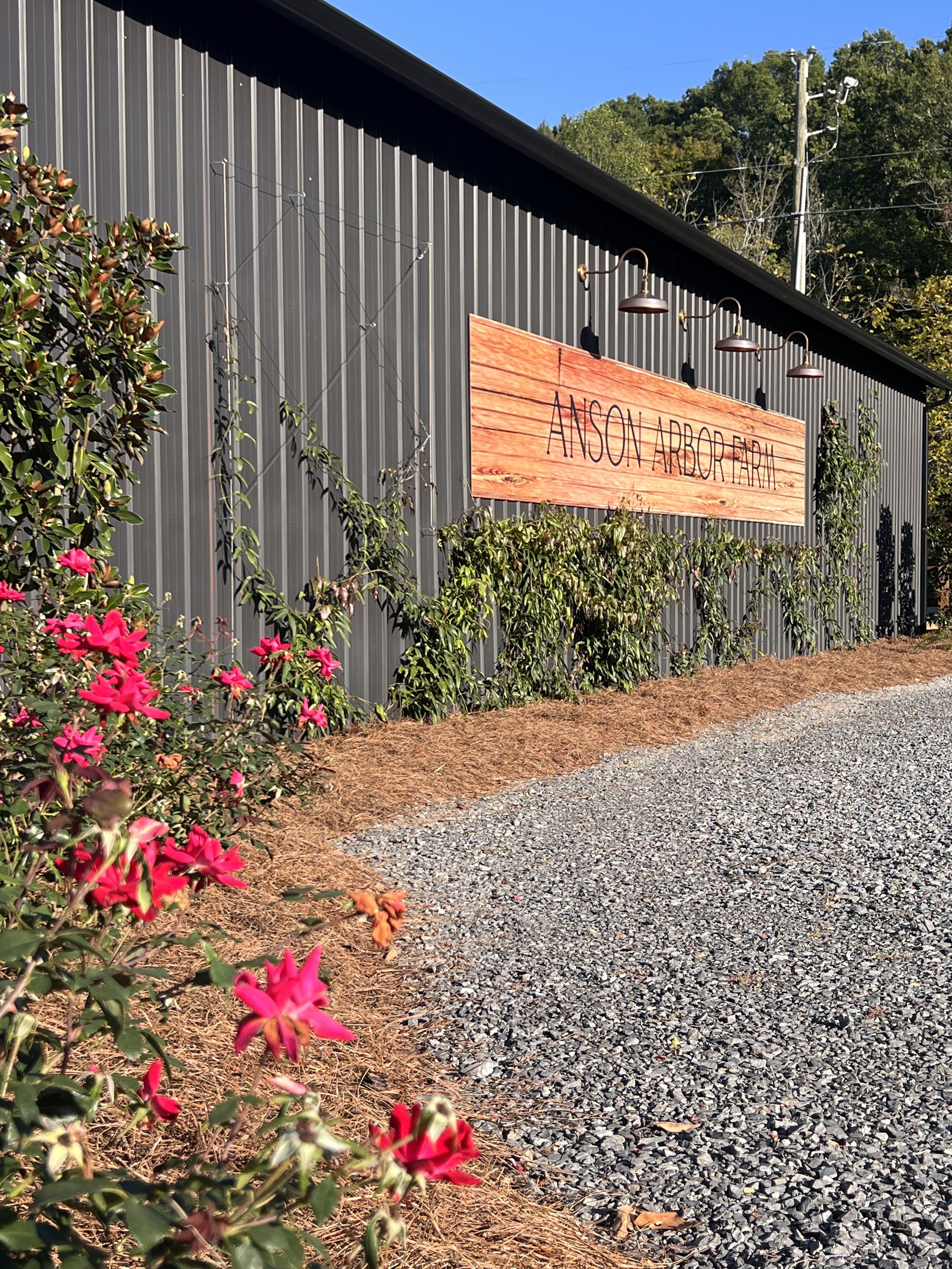 Exterior view of a building with a sign reading 'Anson Arbor Farm,' surrounded by red flowers and greenery, under a clear blue sky.
