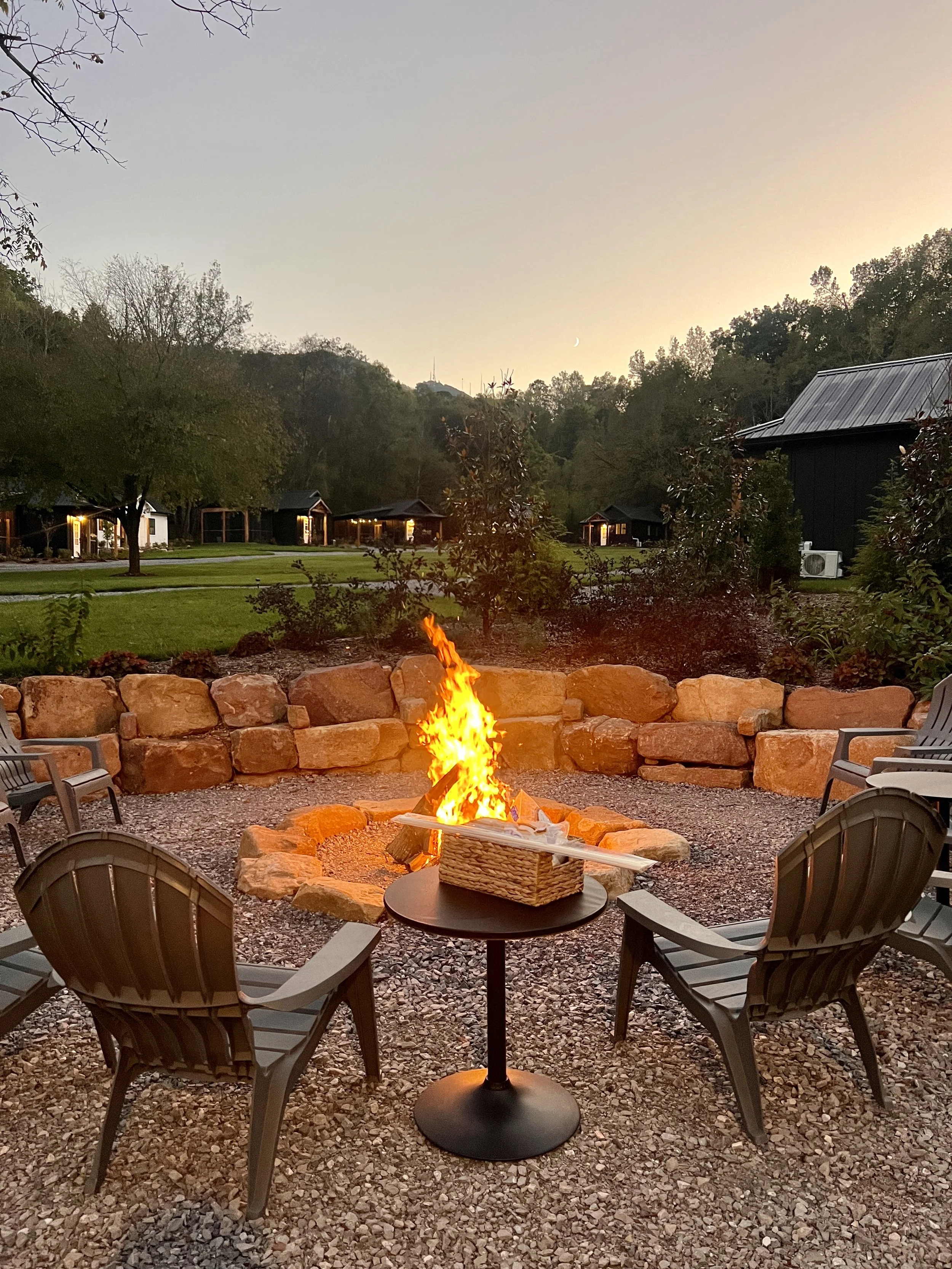 Outdoor fire pit with chairs and table at sunset, surrounded by trees and buildings.