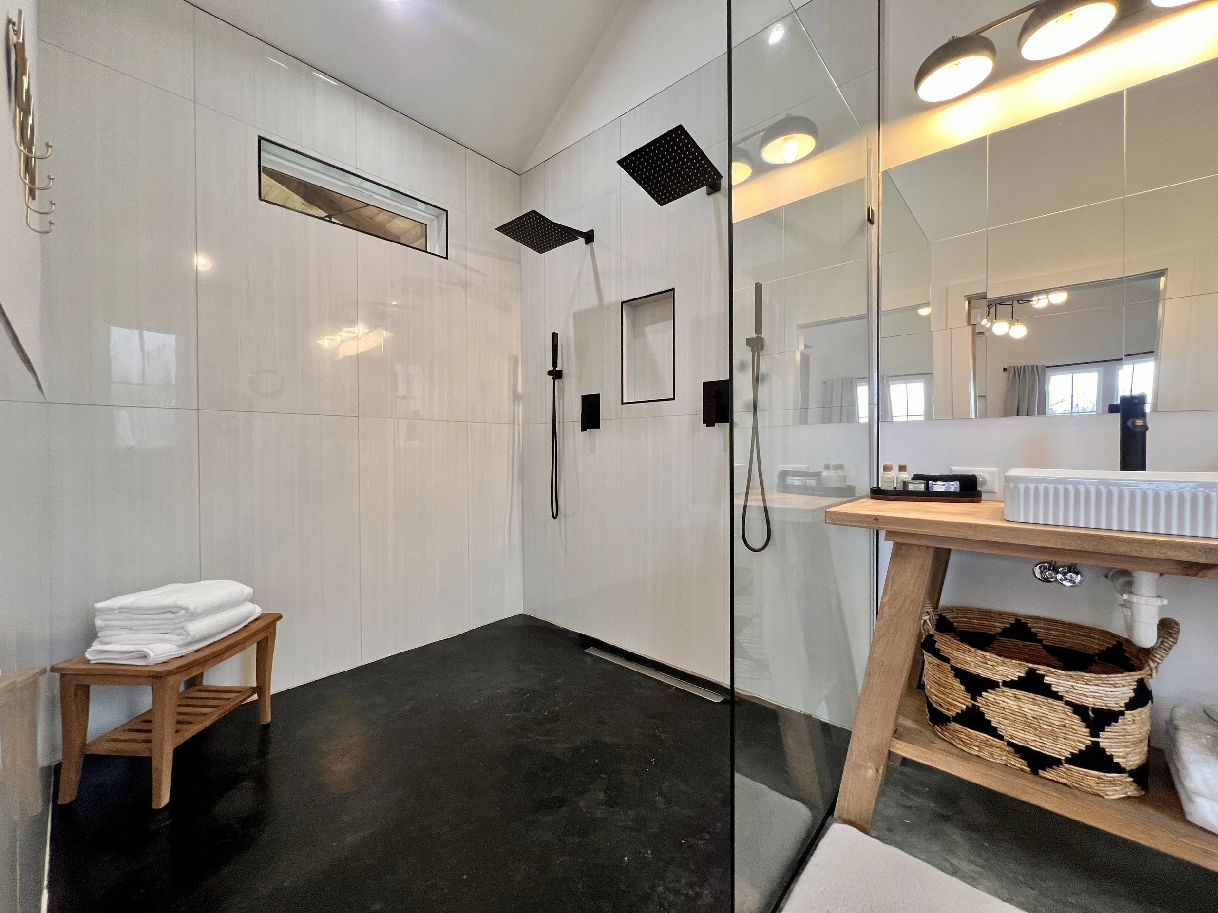 Modern bathroom with a walk-in shower, black floor, wooden bench with white towels, glass partition, vanity with sink, under-sink basket, and lighting fixtures.