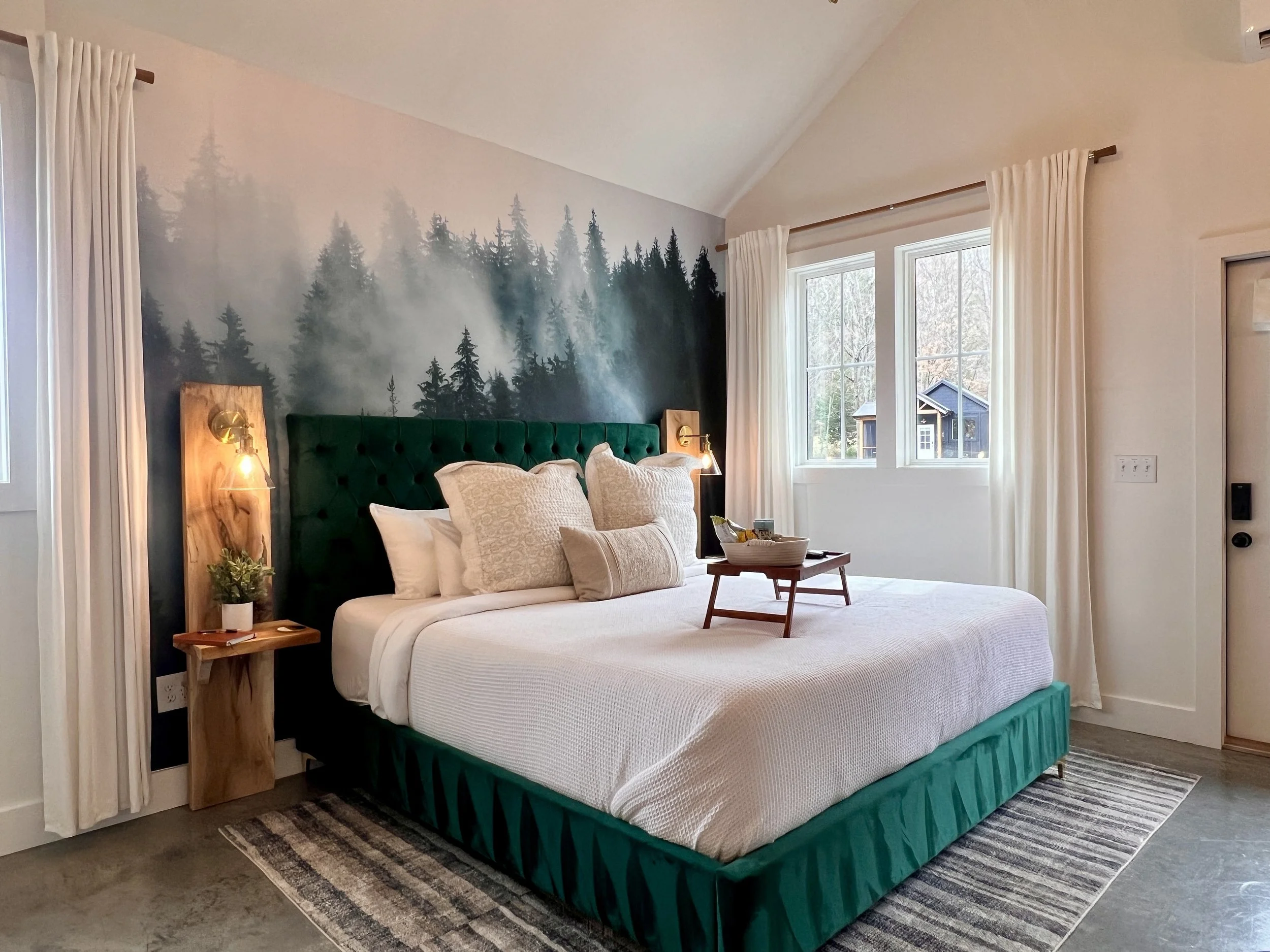 Cozy bedroom with a green upholstered bed and a forest-themed mural on the wall. The room features white curtains, a wooden side table with plants, and a tray on the bed. Large windows provide natural light, and a modern farmhouse style is present th
